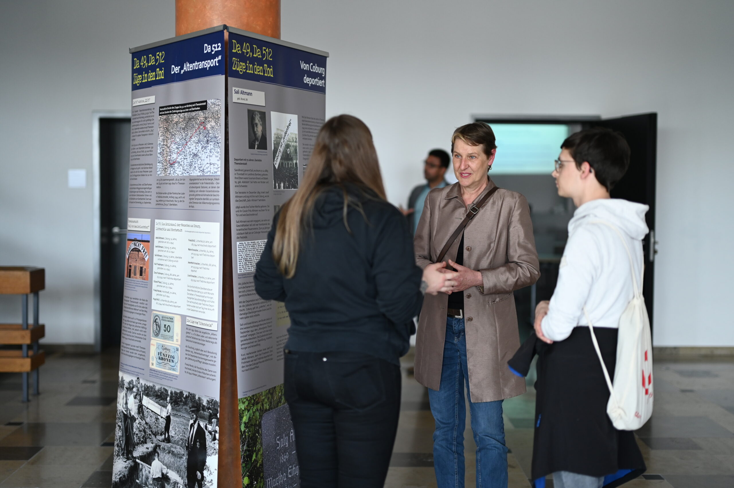 Drei Personen unterhalten sich an einer Informationsausstellung in einem großzügigen Innenraum der Hochschule Coburg. Die Ausstellung zeigt historische Bilder und Texte. Eine Person trägt einen beigen Mantel, eine andere eine dunkle Jacke und die dritte ein weißes Hemd mit einer Tasche.