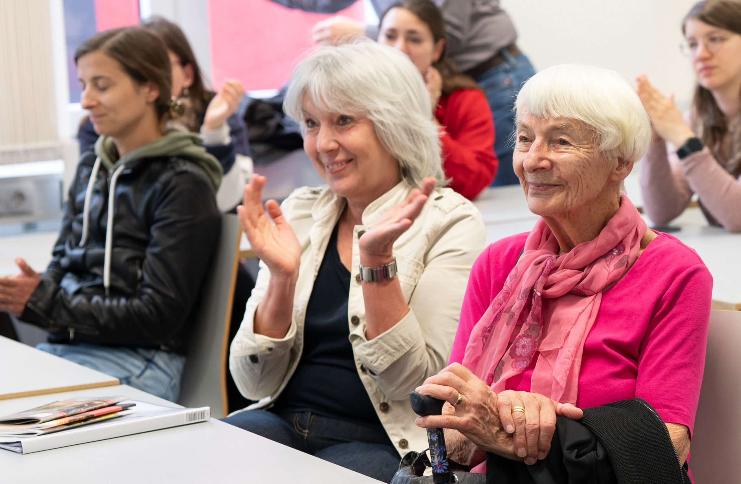 Eine Gruppe von Menschen sitzt in einem Hörsaal der Hochschule Coburg. Im Vordergrund steht eine ältere Frau in einem leuchtend rosa Oberteil und einem Schal, die lächelt. Eine Frau neben ihr klatscht. Andere, leger gekleidet, sitzen im Hintergrund, einige schauen aufmerksam zu.