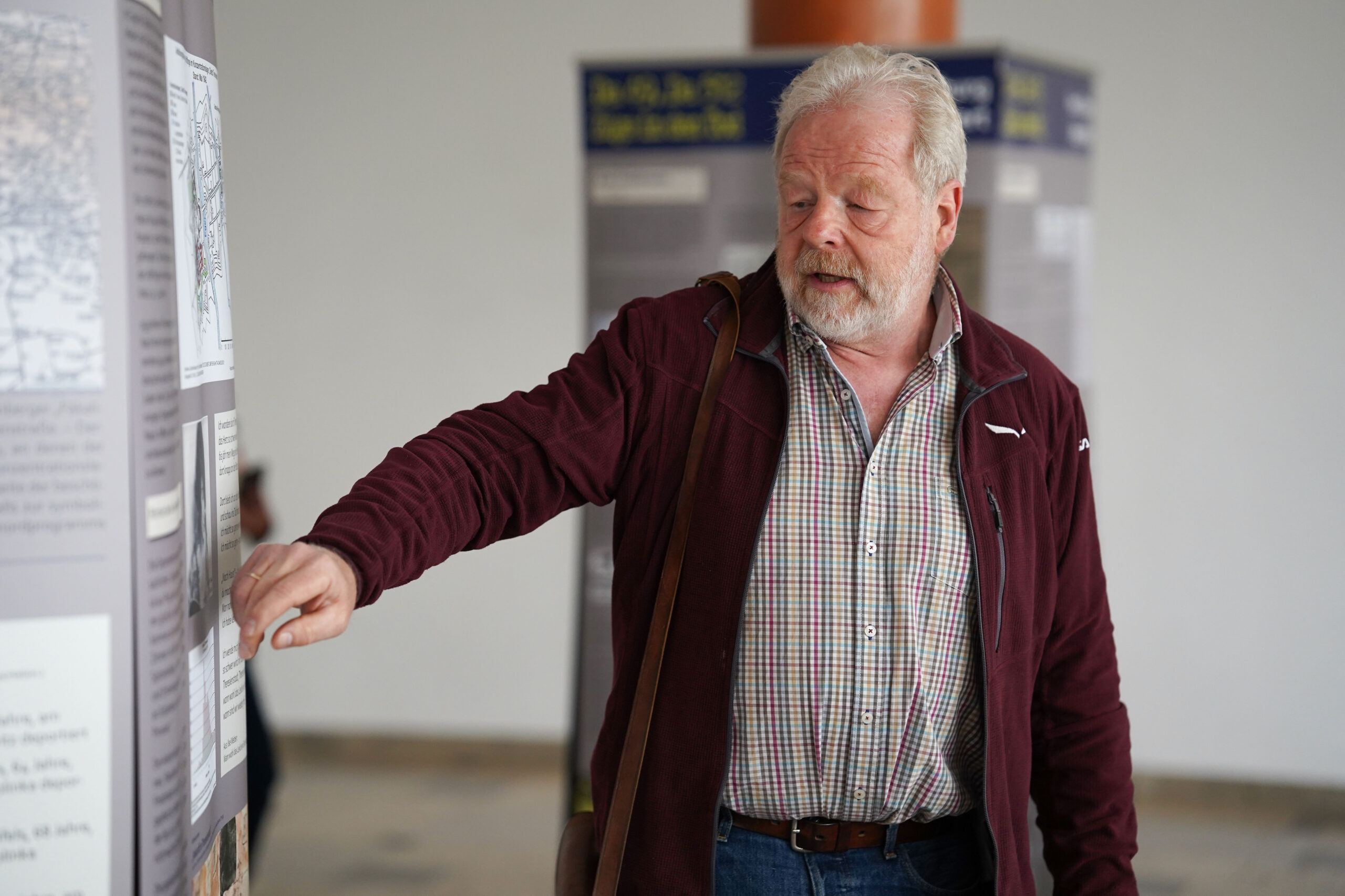 Ein älterer Herr mit Bart, bekleidet mit einem weinroten Jackett und einem karierten Hemd, gestikuliert nachdenklich auf eine Schautafel im Ausstellungsraum der Hochschule Coburg.