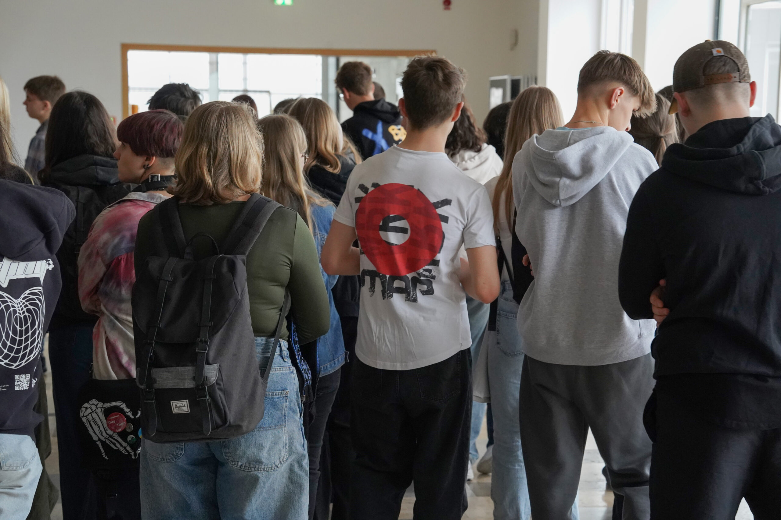 Eine Gruppe von Menschen steht dicht beieinander und blickt abgewandt. Sie tragen legere Kleidung wie T-Shirts, Kapuzenpullover und Rucksäcke und verkörpern damit die entspannte Atmosphäre der Hochschule Coburg. Eine Person trägt in dieser Innenumgebung in der Nähe eines Fensters ein auffälliges T-Shirt mit einem roten Kreismuster.