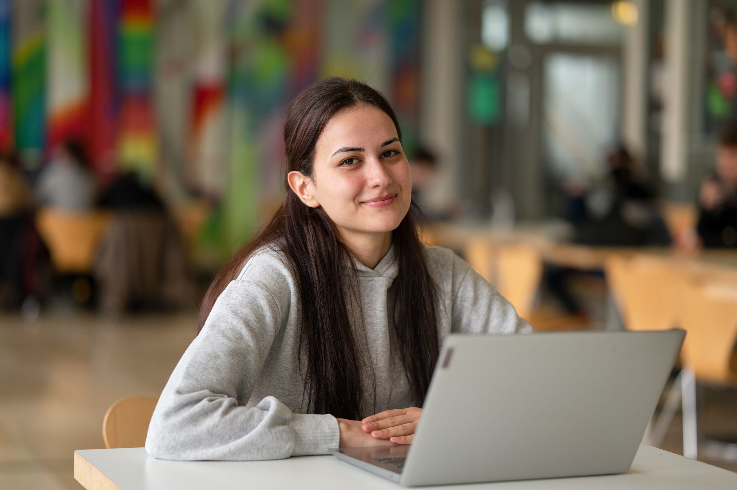 Eine Person mit langen dunklen Haaren sitzt lächelnd an einem Tisch, vor ihr liegt ein aufgeklappter Laptop. Die lebhafte Kulisse lässt vermuten, dass es sich um die Hochschule Coburg handelt. Hier wimmelt es von Studierenden und Lehrkräften, die an verschiedenen Tischen im Raum lebhafte Diskussionen führen.