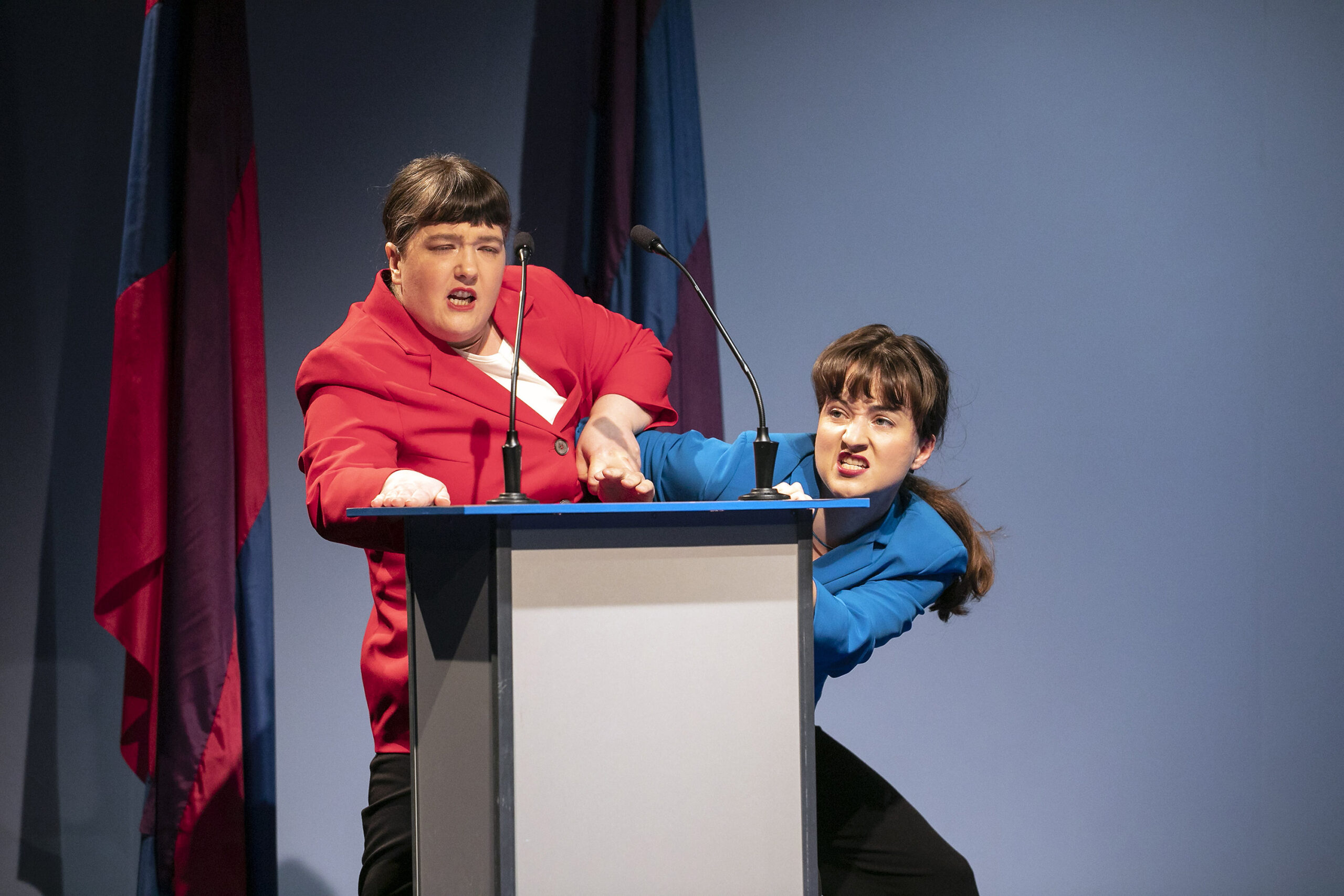 Zwei Personen stehen auf der Bühne vor einem Podium mit Mikrofonen und repräsentieren die Hochschule Coburg. Einer in einer roten Jacke hält den Arm des anderen in einer blauen Jacke. Vor dem Hintergrund der rot-blauen Fahnen wirken ihre Ausdrücke intensiv.