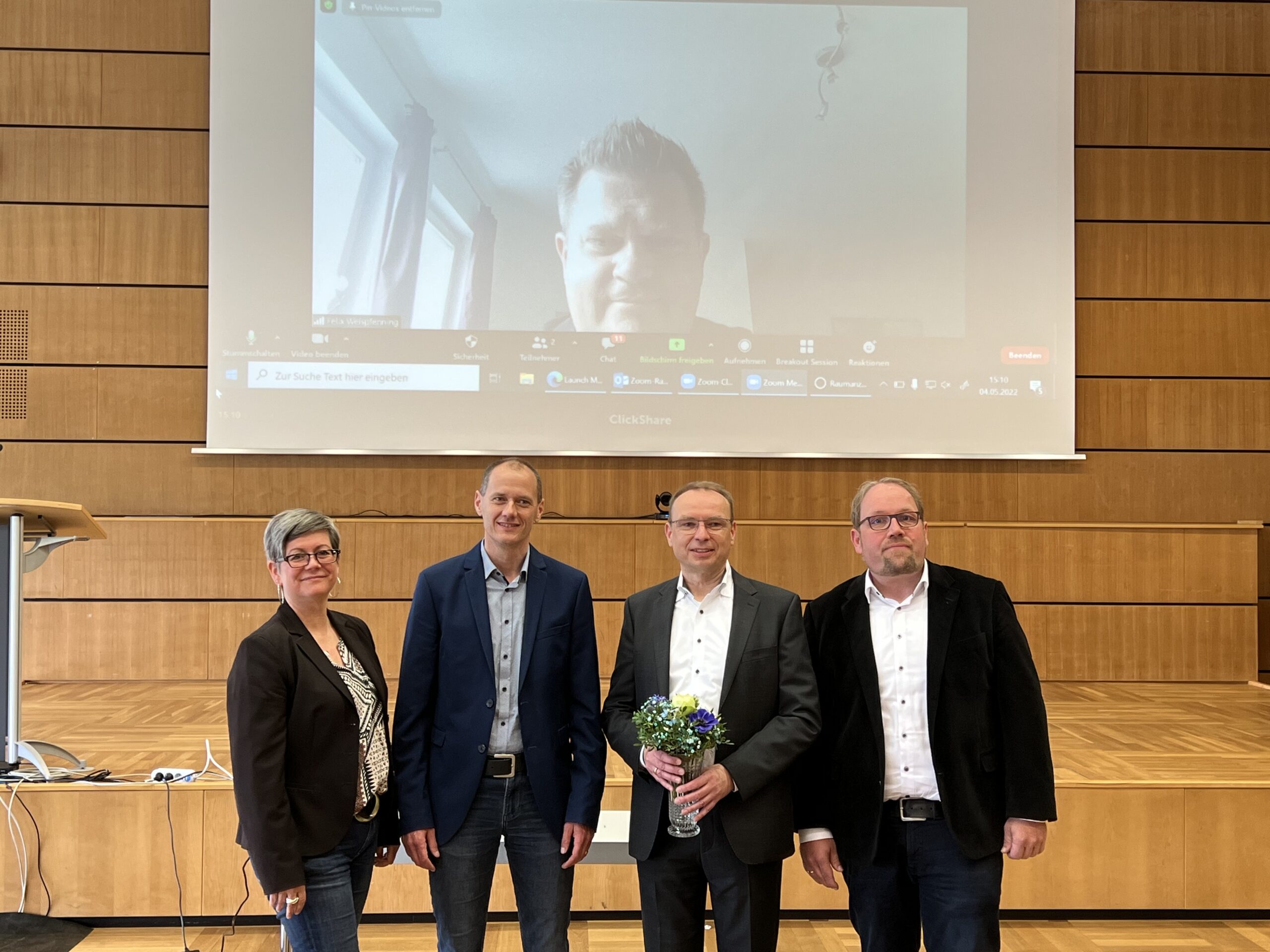 Vier Menschen stehen in einem Raum mit Holzwänden und verkörpern den Geist der Hochschule Coburg. Eine Person hält Blumen in den Händen, die den Feierlichkeiten ein Symbol sind. Auf einem großen Bildschirm hinter ihnen ist eine weitere Person zu sehen, die per Videoanruf an der Versammlung teilnimmt.