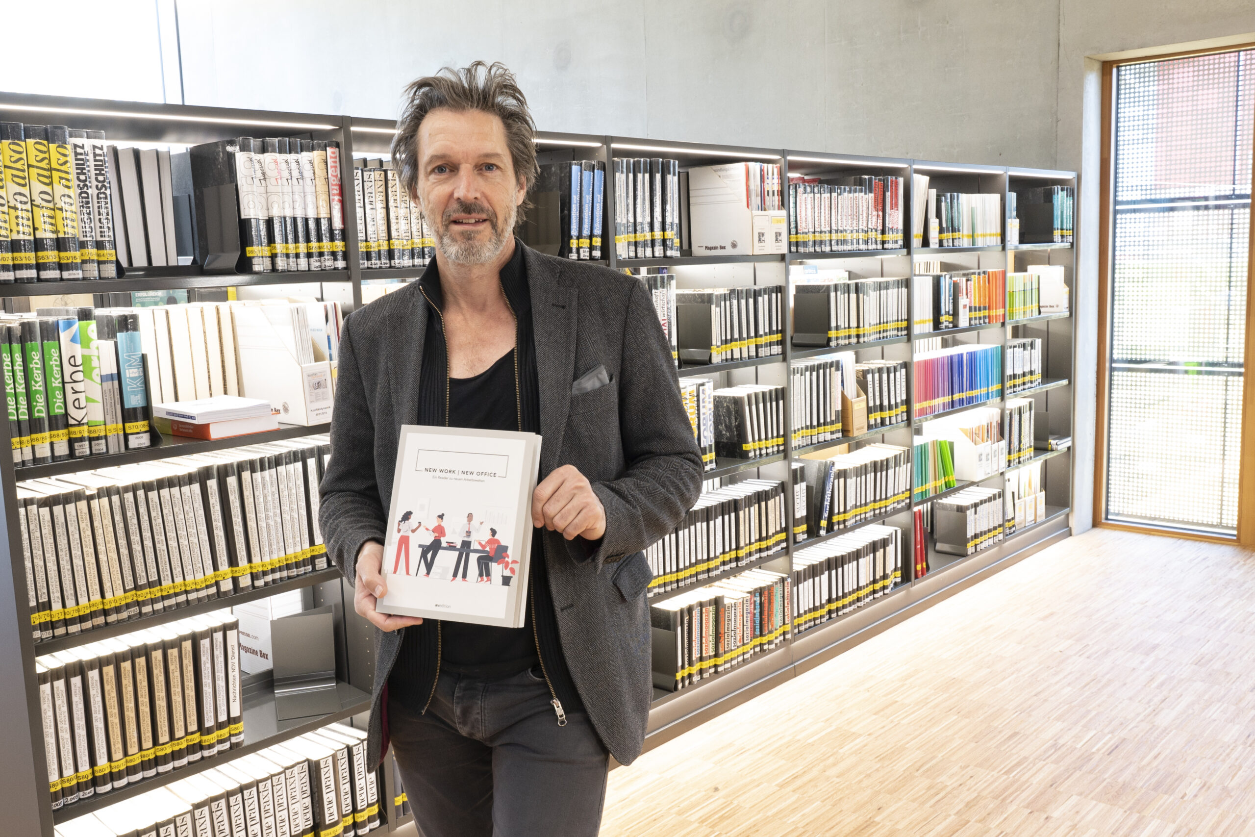 Ein Mann mit braunen Haaren und grauem Bart steht vor den Bücherregalen der Hochschule Coburg und hält ein Buch mit dem Titel „50 Jahre KHM: MAMIT“ in der Hand. Die Regale sind ordentlich mit Büchern gefüllt, in einem hellen Raum mit großem Fenster.