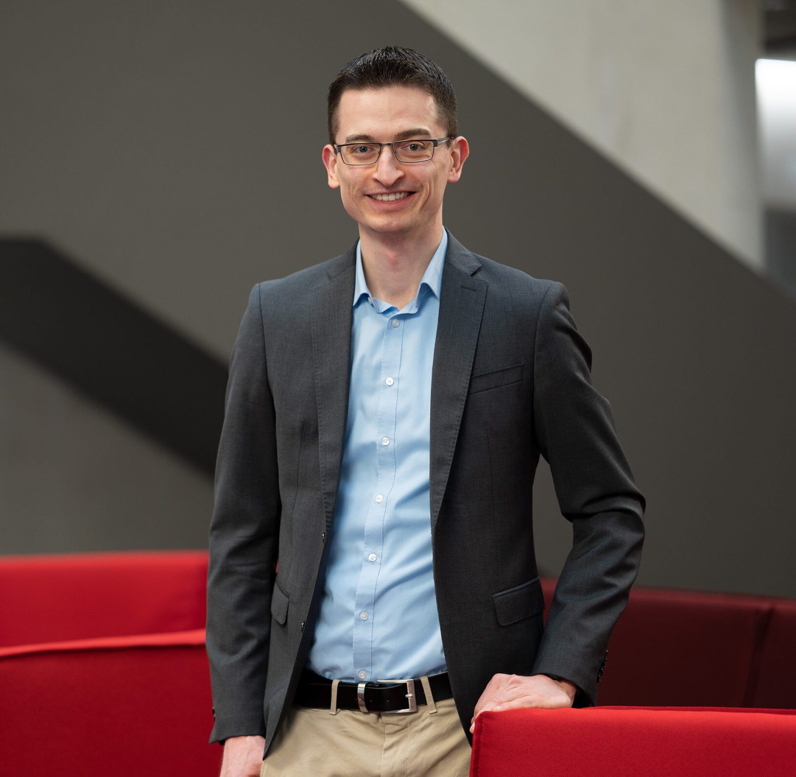 Eine Person mit Brille, grauem Blazer, hellblauem Hemd und Khakihose steht lächelnd neben einem roten Sofa an der Hochschule Coburg. Im Hintergrund ist eine graue Wand zu sehen.