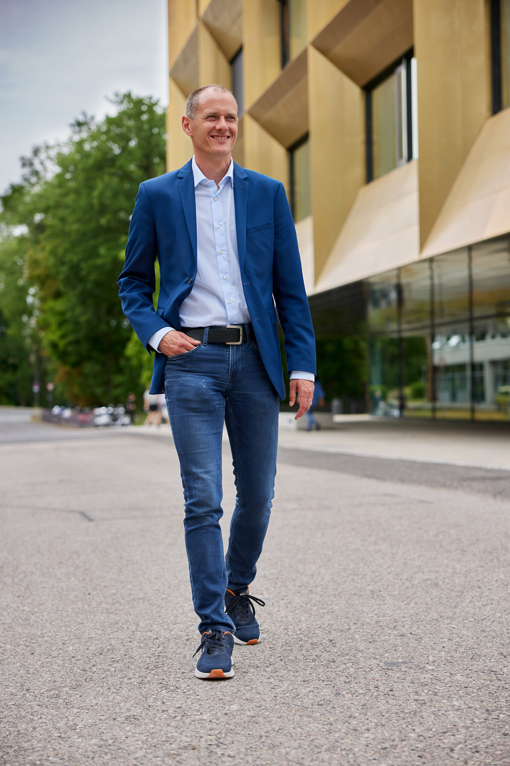 Ein Mann in blauem Blazer und Jeans schreitet selbstbewusst eine Stadtstraße entlang. Die moderne Kulisse mit großen Fenstern und von Bäumen gesäumten Gehwegen erinnert an den lebendigen Campus der Hochschule Coburg. Er lächelt und strahlt Entspannung und Gelassenheit aus.