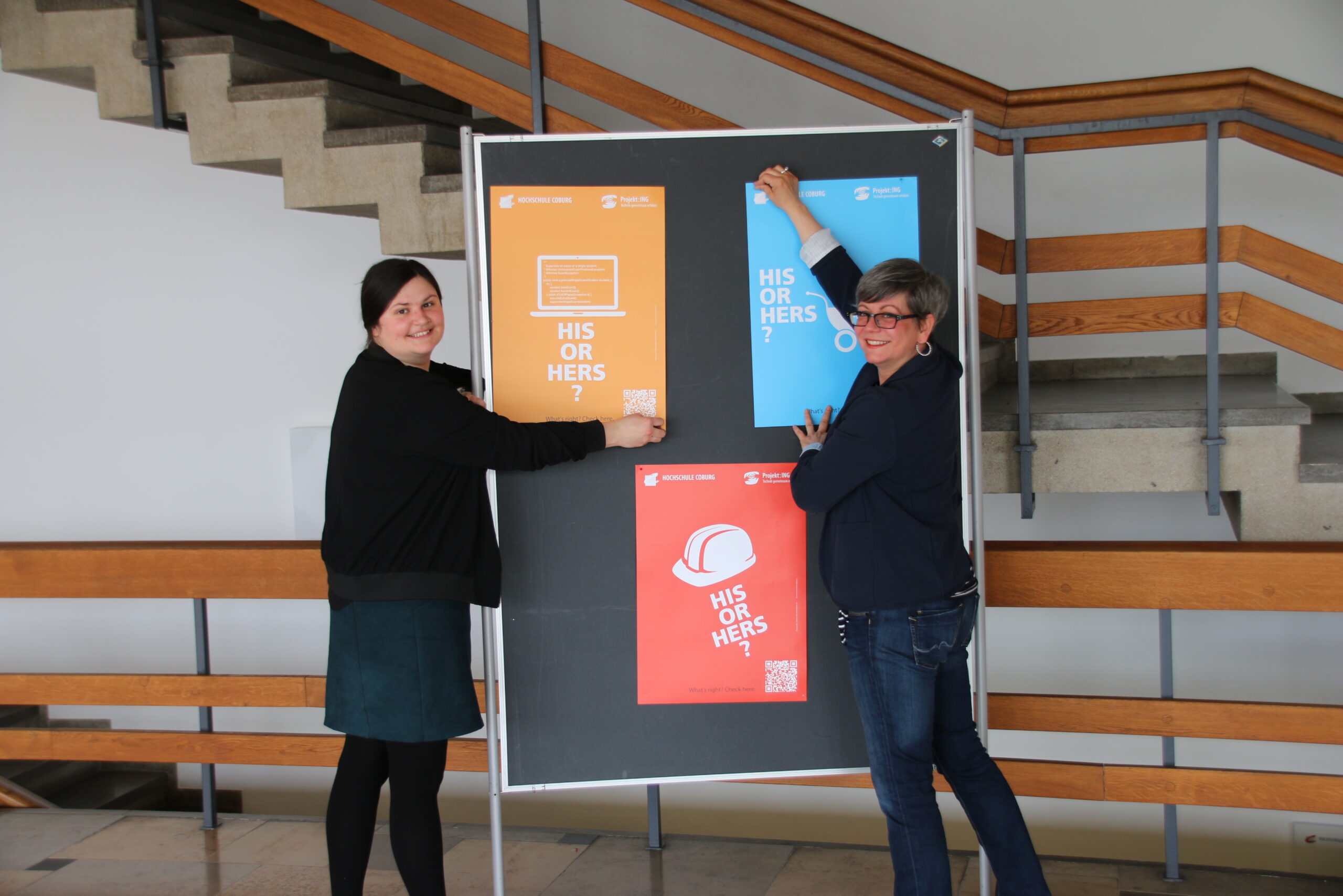 Zwei Personen hängen bunte Plakate an ein schwarzes Brett der Hochschule Coburg. Die mit Text und Bildern gefüllten Plakate stehen vor einer Treppe mit Holzgeländer in einem hellen Raum. Sie lächeln in die Kamera und fangen die lebendige Atmosphäre des Campus ein.