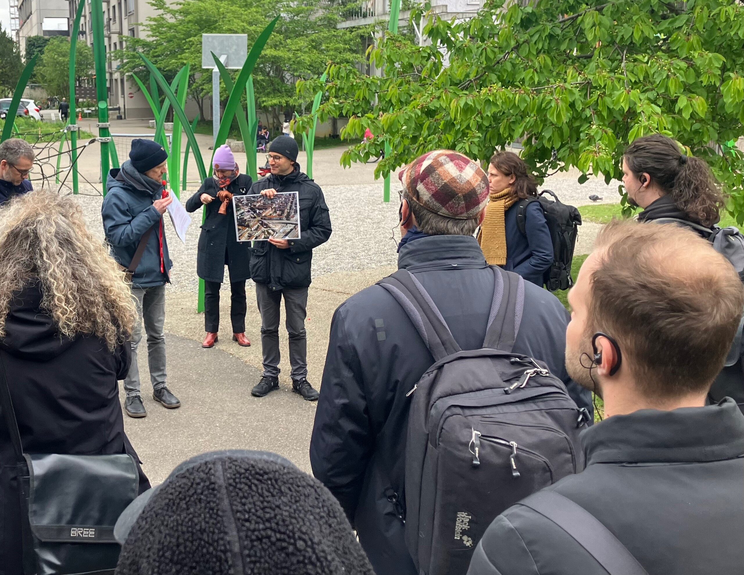 A group of people gathered outdoors, listening to a speaker from Hochschule Coburg holding a large photo. They are dressed in jackets and hats, suggesting cool weather. The background features tall green structures and trees.