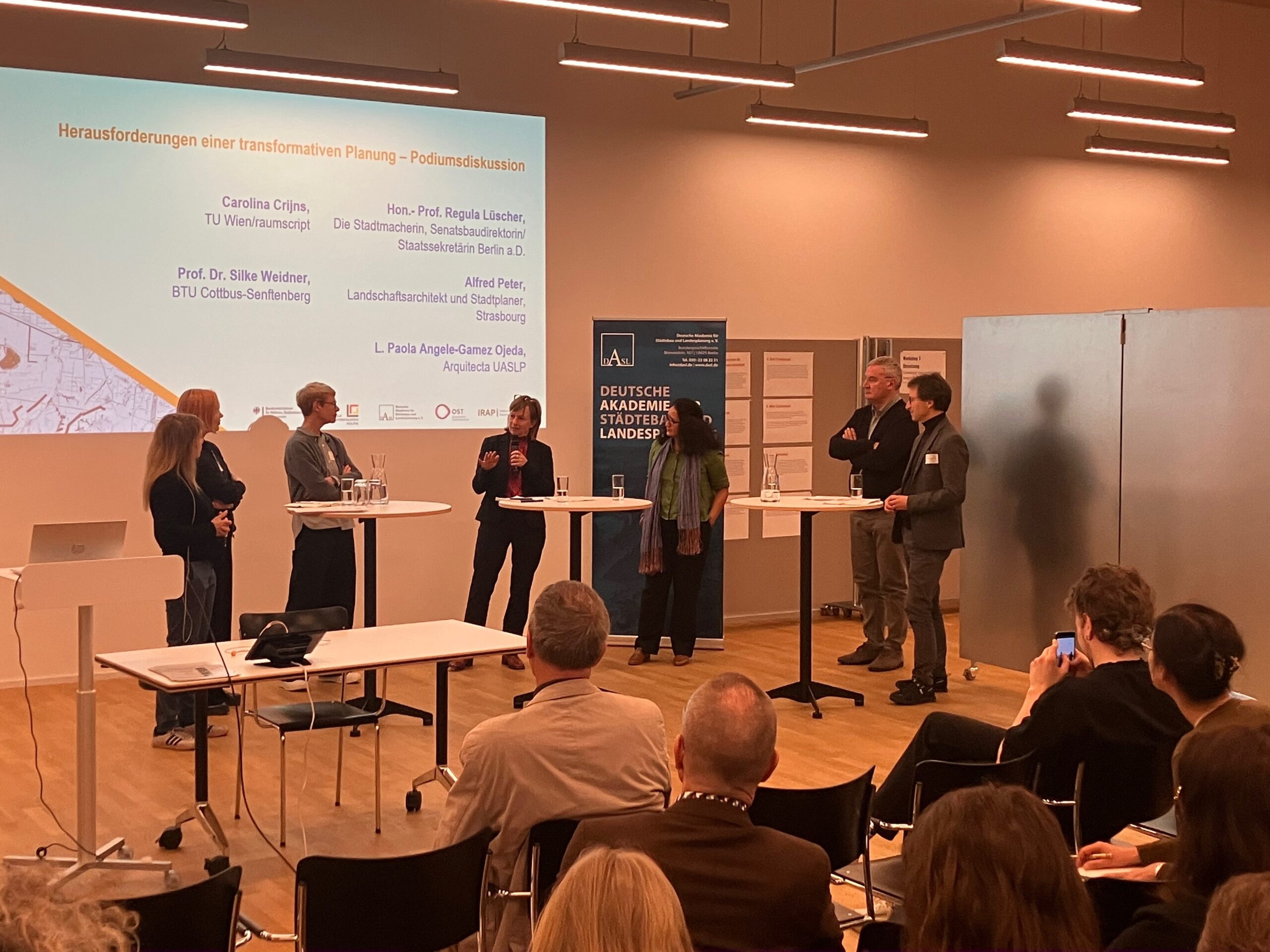 A group of six people stands in a semi-circle, engaging in a panel discussion at Hochschule Coburg's conference room. A presentation is displayed on a screen behind them. An audience is seated in the foreground, listening attentively.