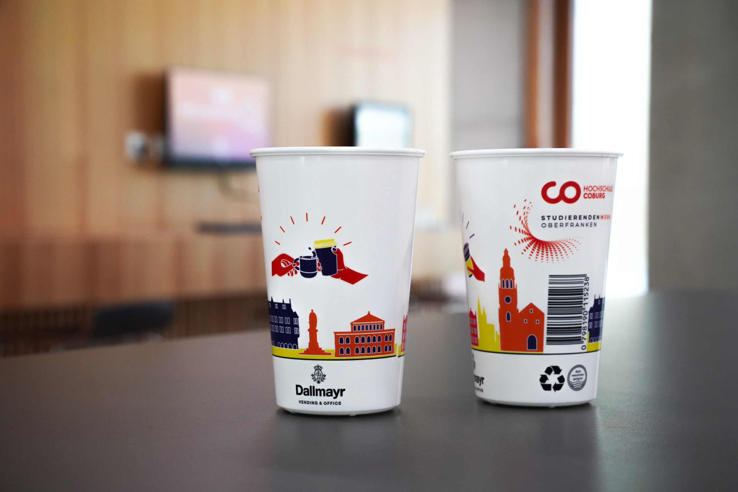 Two white cups adorned with colorful city-themed illustrations and logos rest on the table. In the blurred background, a wooden wall and a TV screen subtly frame the scene, reminiscent of a cozy Hochschule Coburg café ambiance.