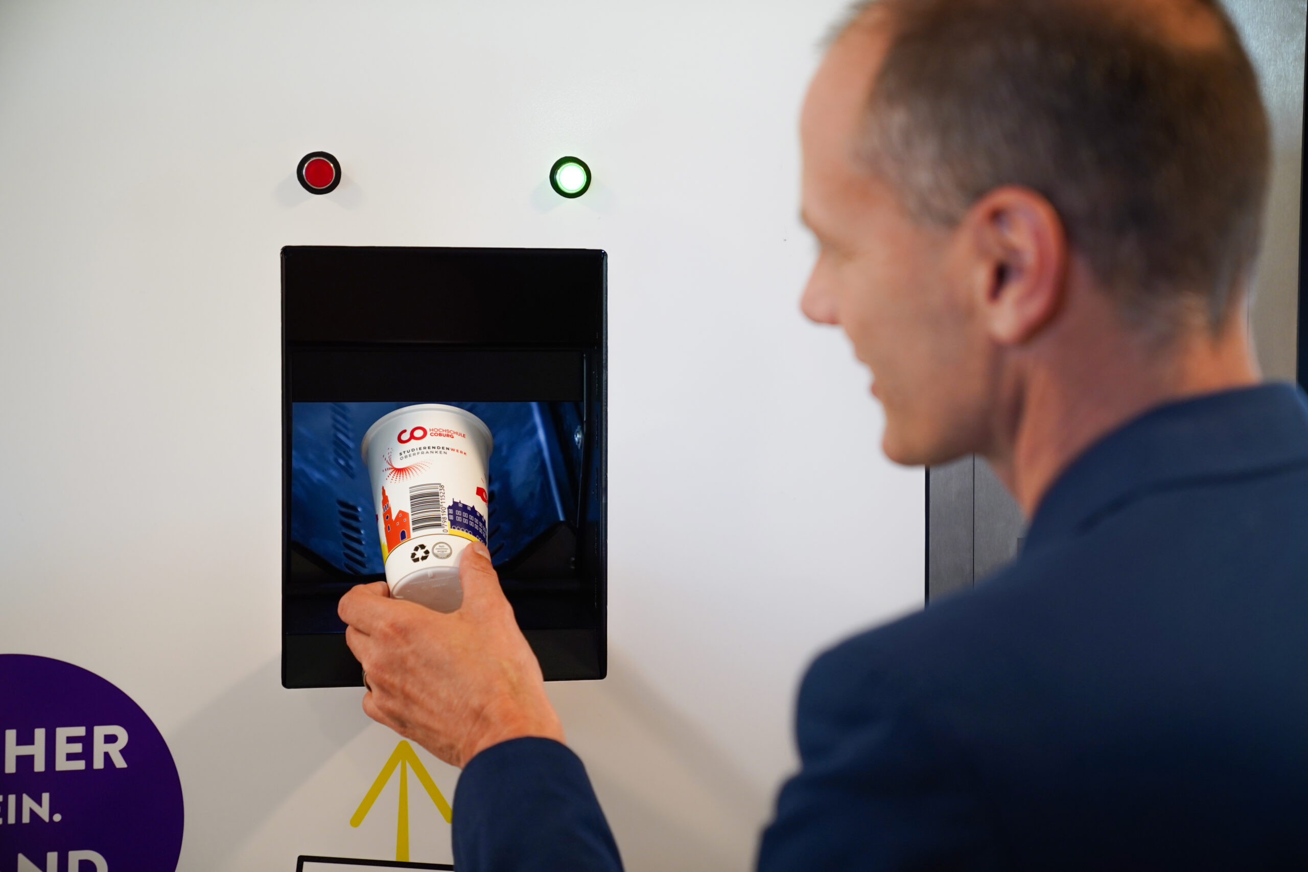 A person inserts a white cup with red and blue Hochschule Coburg branding into a recycling machine with a rectangular slot. The machine displays a green light, indicating acceptance. The person's hand holds the cup, and their face is slightly turned away.