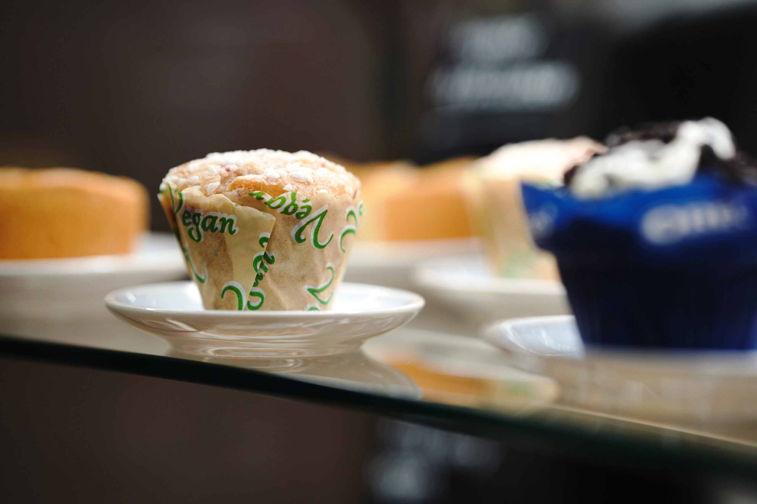 A vegan muffin on a small white plate, wrapped in green paper with "vegan" written on it, is proudly displayed on a glass shelf. In the background at Hochschule Coburg’s café, other baked goods appear enticingly blurred.