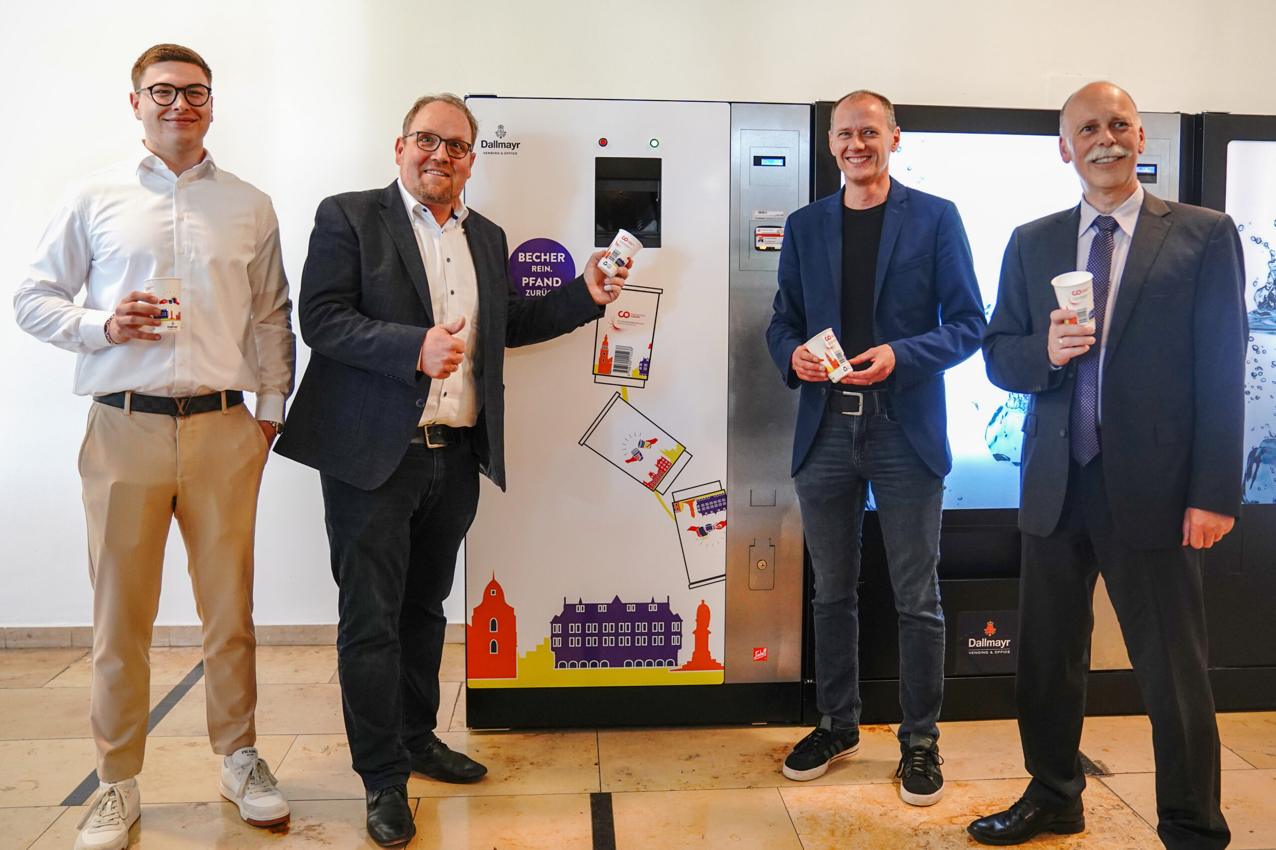 Four men in formal attire stand in front of a colorful vending machine at Hochschule Coburg. Each holding cups and smiling broadly, they give thumbs up amid vibrant images of buildings. The indoor setting with its tiled floor adds to the cheerful atmosphere.