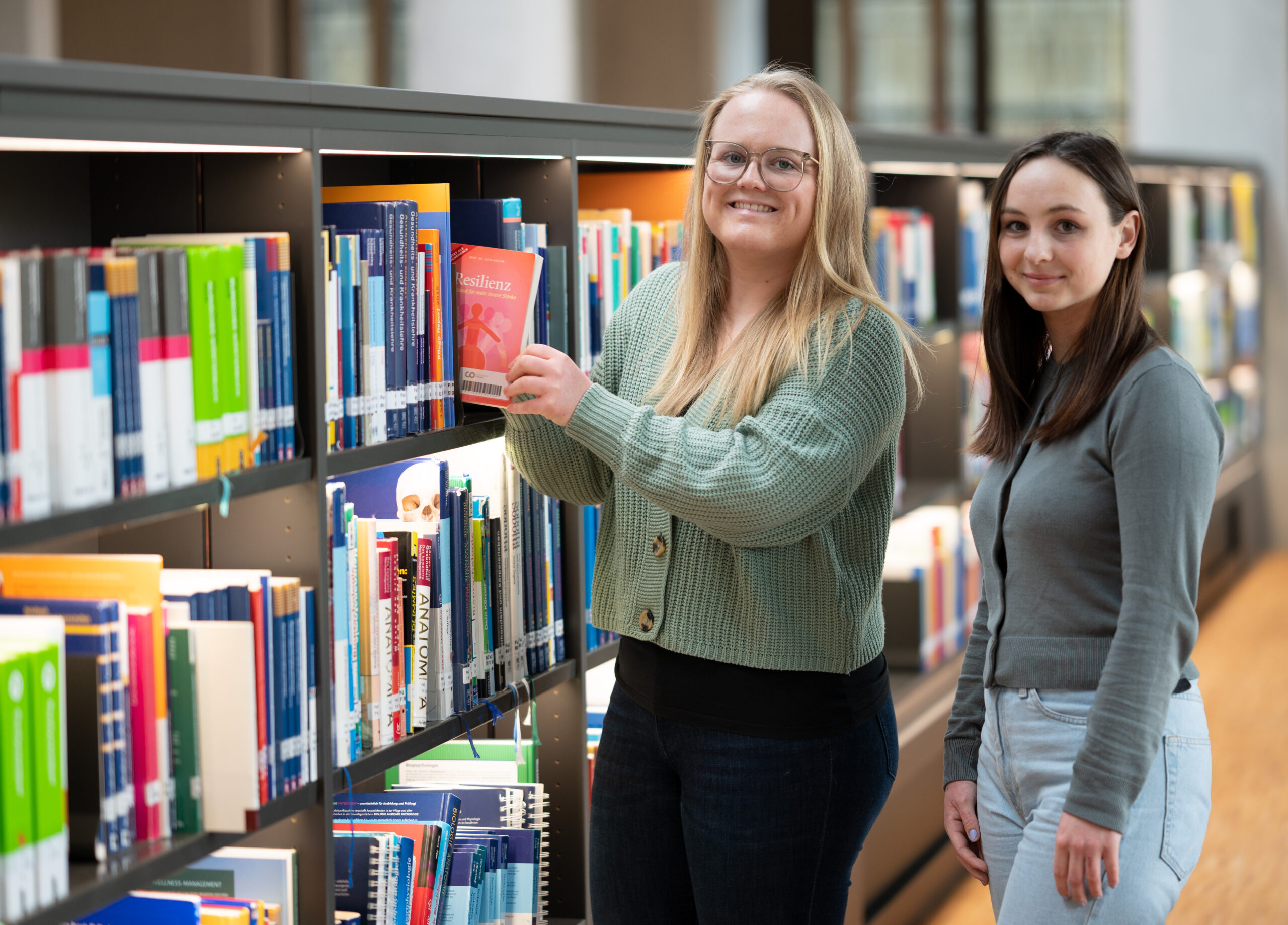 Zwei Frauen stehen vor den Bücherregalen der Bibliothek der Hochschule Coburg. Eine Frau mit Brille sucht sich ein Buch aus, die andere lächelt in die Kamera. Die Bibliothek ist gut beleuchtet, die Regale sind mit bunten Büchern gefüllt.