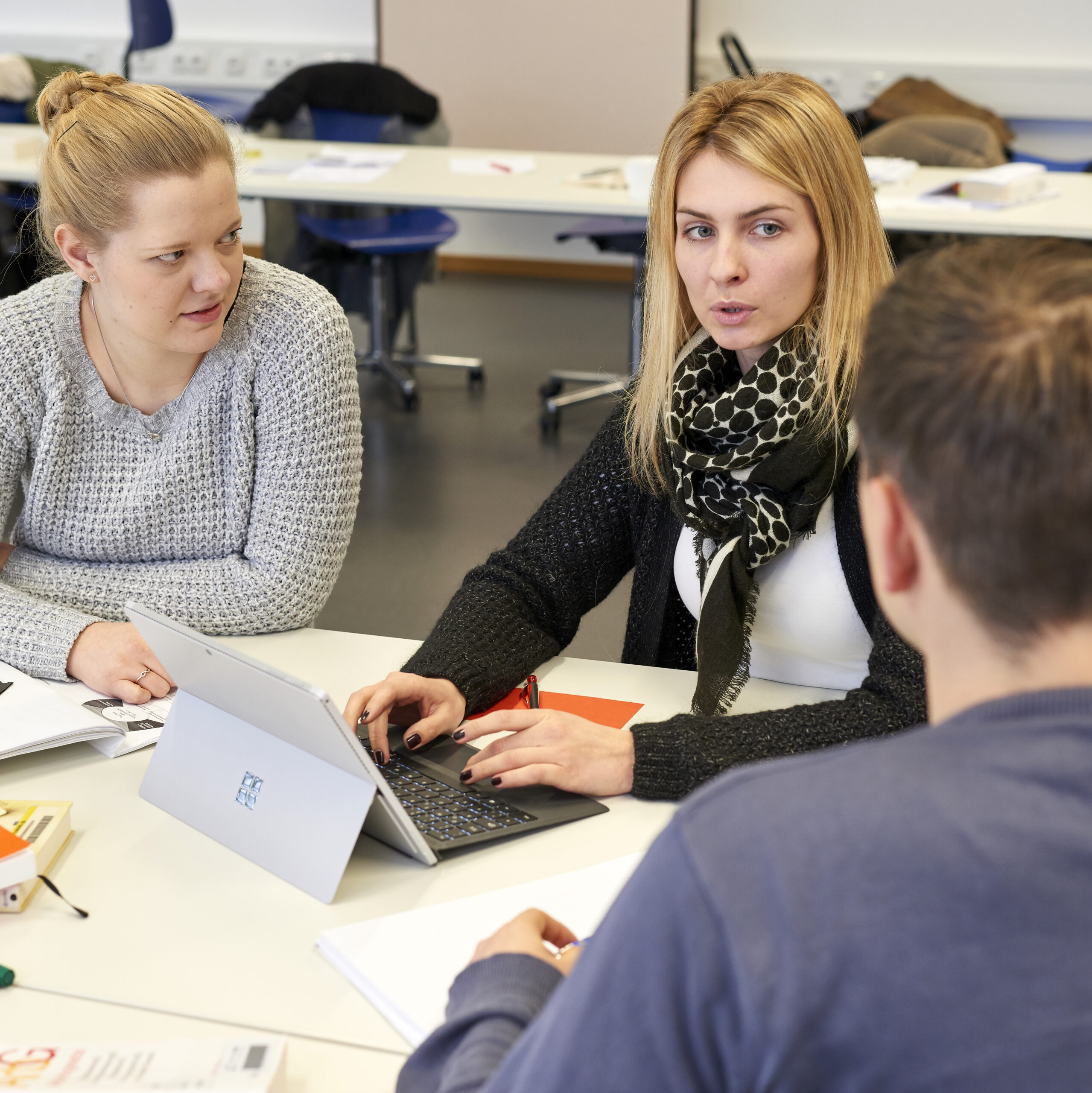 Drei Personen sitzen an einem Tisch in einem Hörsaal der Hochschule Coburg und diskutieren. Eine Person benutzt einen Laptop, auf dem Tisch liegen Papiere und Notizbücher. Der Raum hat weiße Wände und im Hintergrund Schreibtische.