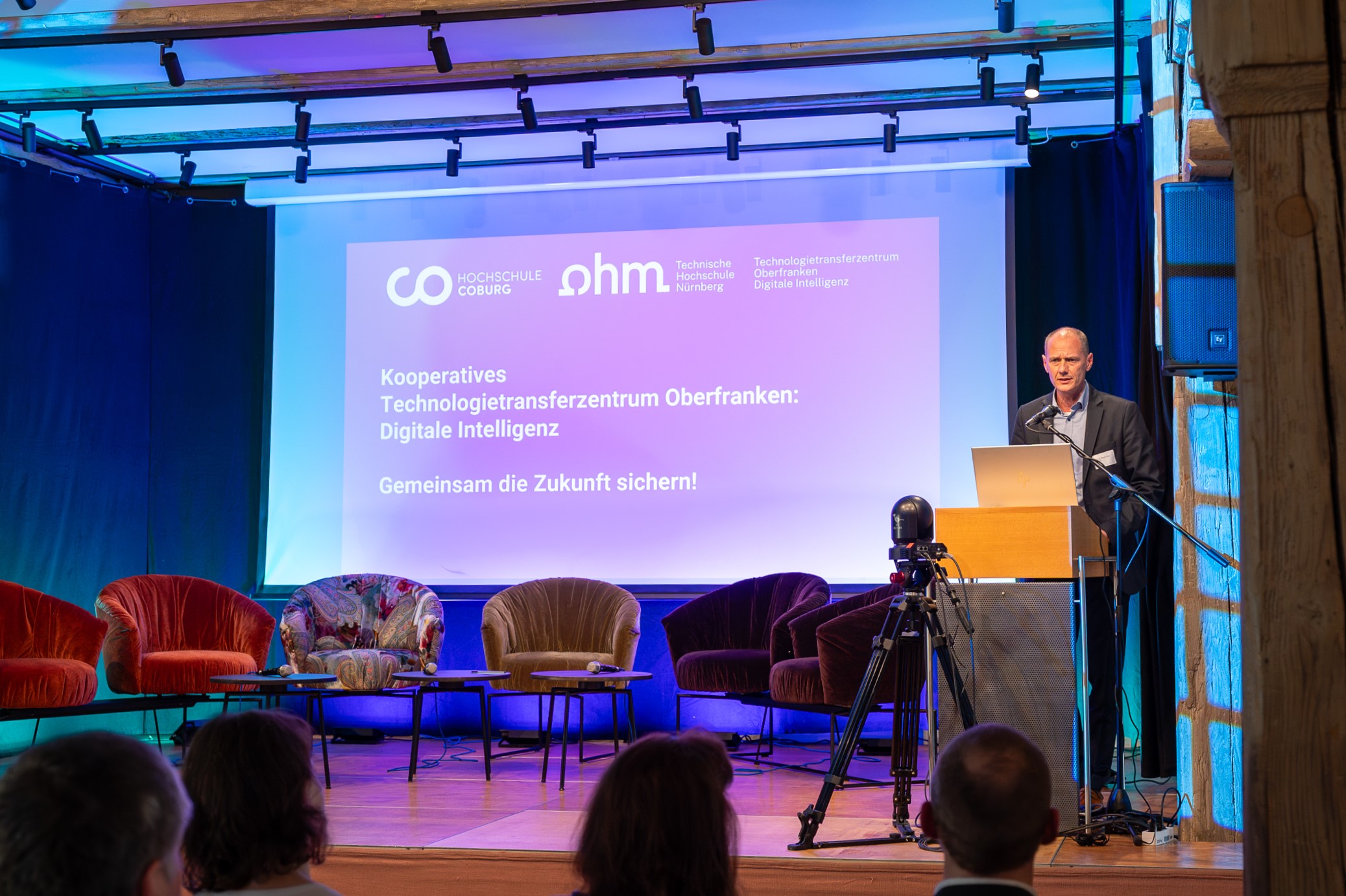 A speaker stands at a podium on a stage at Hochschule Coburg, presenting in front of a blue-lit screen with text about digital intelligence and collaboration. Empty colorful chairs line the stage, and an audience is visible in the foreground.