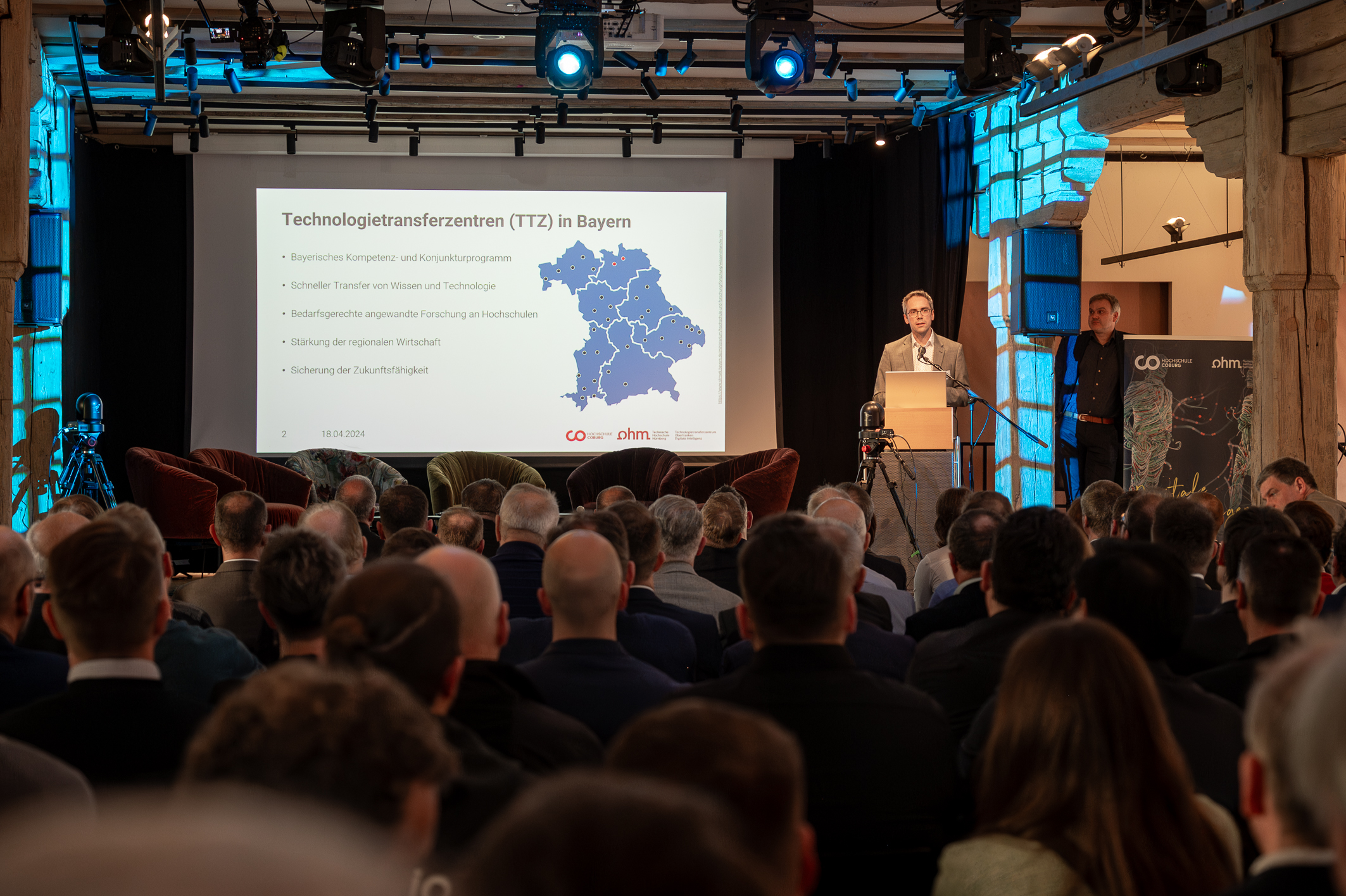 A conference room filled with seated attendees. A speaker from Hochschule Coburg is presenting at a podium next to a large screen showing a map of Bavaria with text about technology centers. The room features spotlights and exposed brick walls, creating an engaging atmosphere.