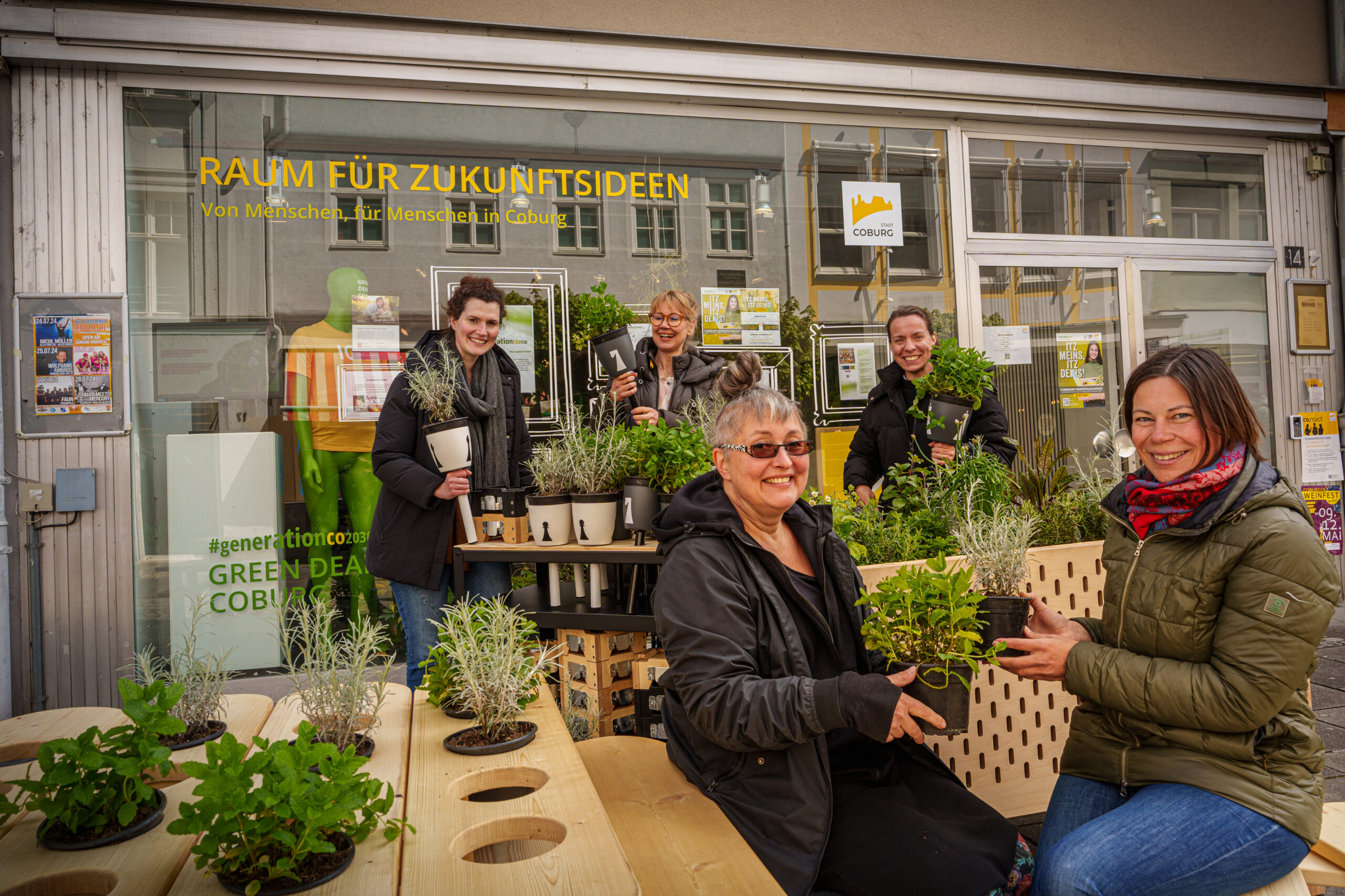 A group of five people are happily gathered in front of a shop with large windows, reminiscent of the Hochschule Coburg community spirit. They are arranging various potted plants on a wooden table. The shop sign reads "Raum für Zukunftsideen.