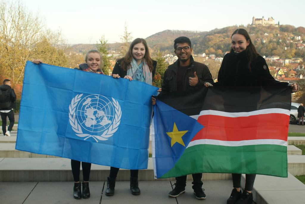 Vier Menschen lächeln und halten Flaggen im Freien: Einer hält eine Flagge der Vereinten Nationen, während andere stolz die Flagge des Südsudan zeigen. Vor der malerischen Kulisse von Hügeln und Gebäuden feiern diese Studenten der Hochschule Coburg Einheit und globale Bildung.