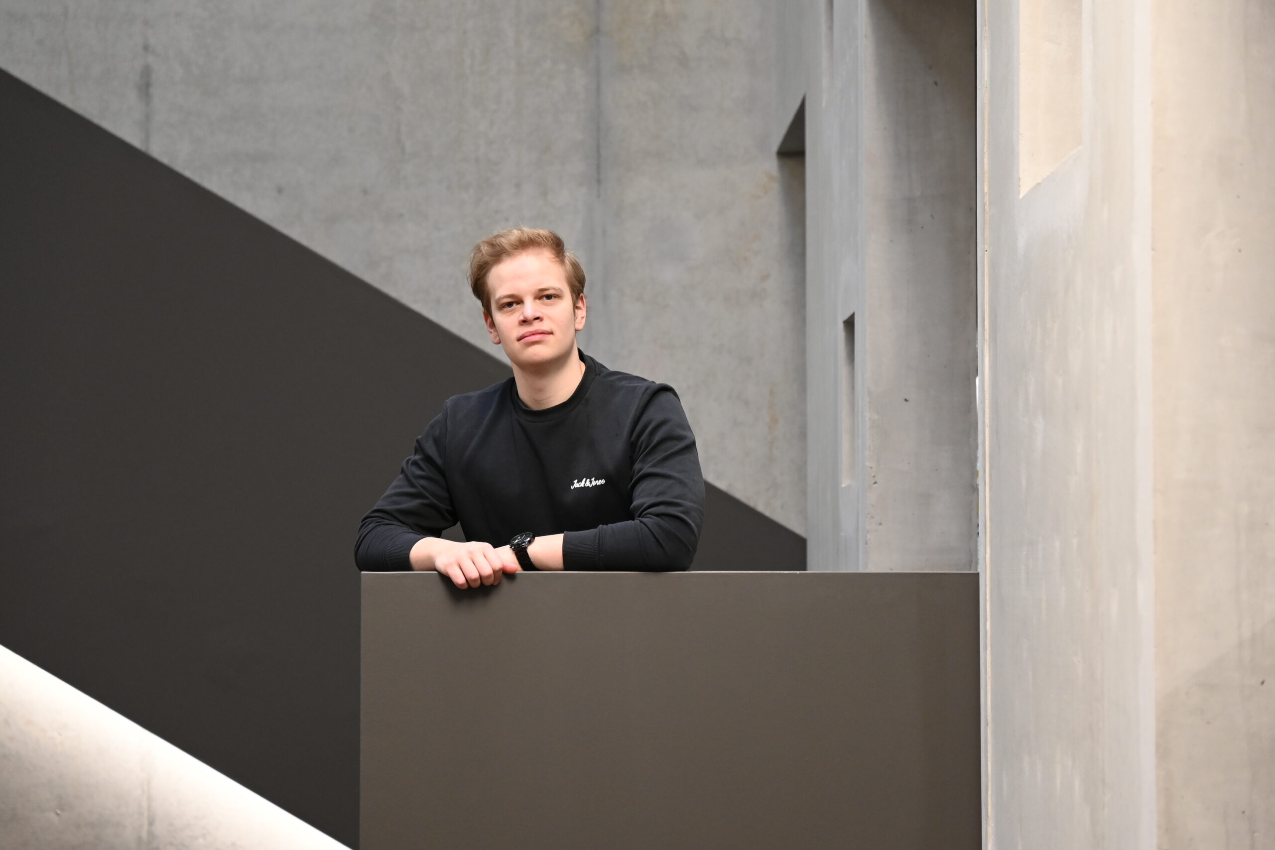 Eine Person mit dunklem Pullover und Uhr lehnt an einem grauen Geländer der Hochschule Coburg. Im Hintergrund ist eine moderne, minimalistische Architektur mit Betonwänden und geometrischen Formen zu sehen.