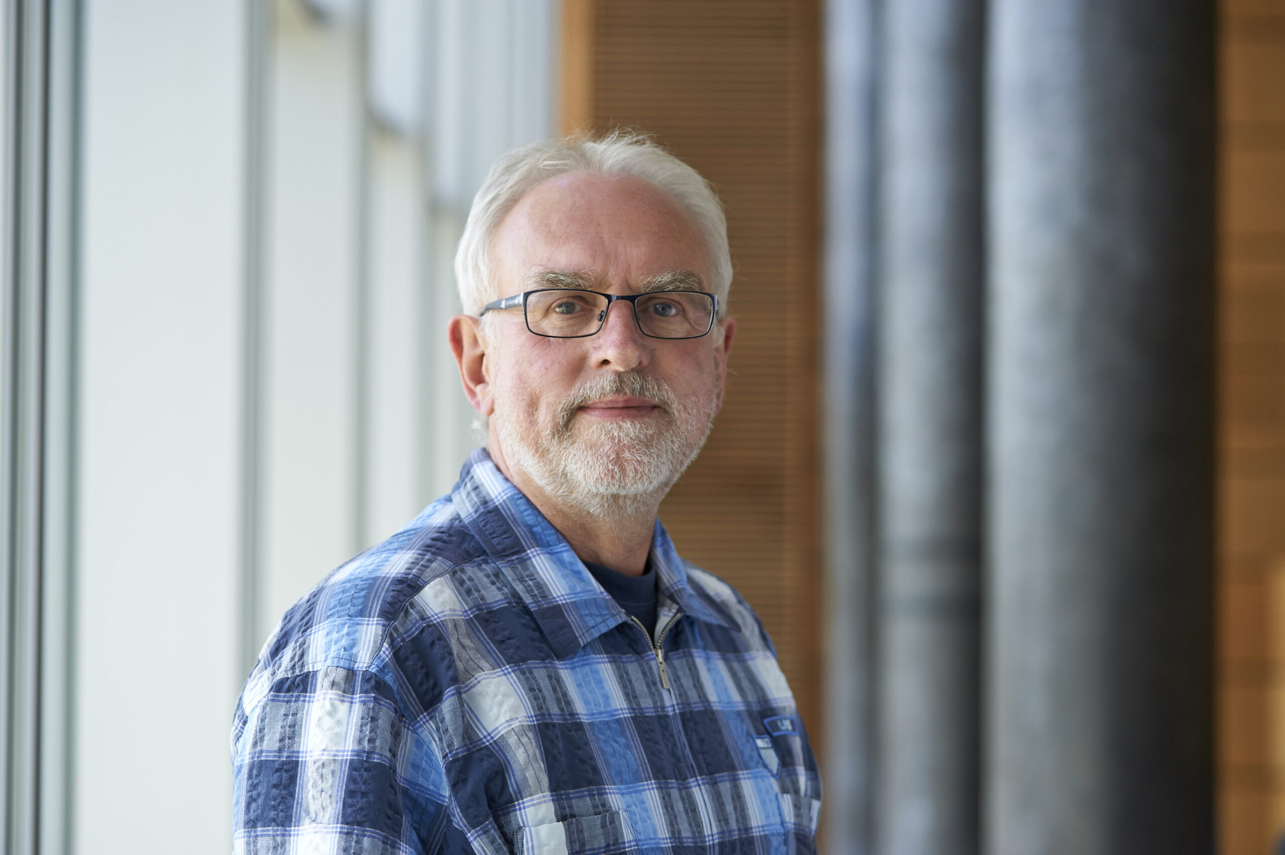 Ein älterer Mann mit Brille und Bart steht drinnen und trägt ein blau kariertes Hemd. Er blickt mit neutralem Gesichtsausdruck nach vorne. Die moderne Inneneinrichtung, die dem Design der Hochschule Coburg ähnelt, zeichnet sich durch große Fenster und Holzpaneele aus.