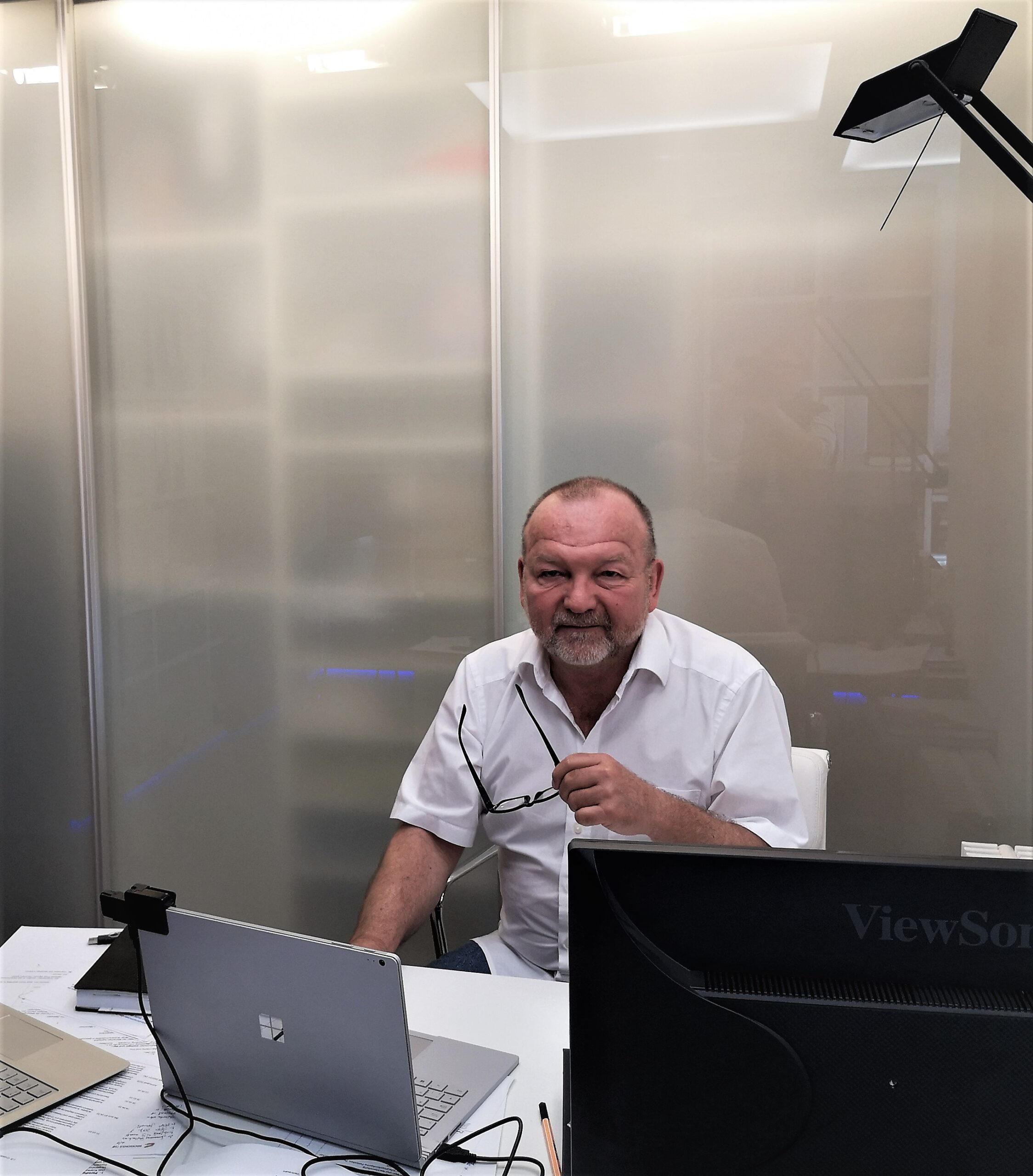 Ein Mann mit Bart und weißem Hemd sitzt an einem Schreibtisch vor zwei Computermonitoren. Er hält eine Brille in der Hand und lächelt. Im Hintergrund sind Milchglasscheiben zu sehen, die eine akademische Atmosphäre schaffen, die an die modernen Studienräume der Hochschule Coburg erinnert.