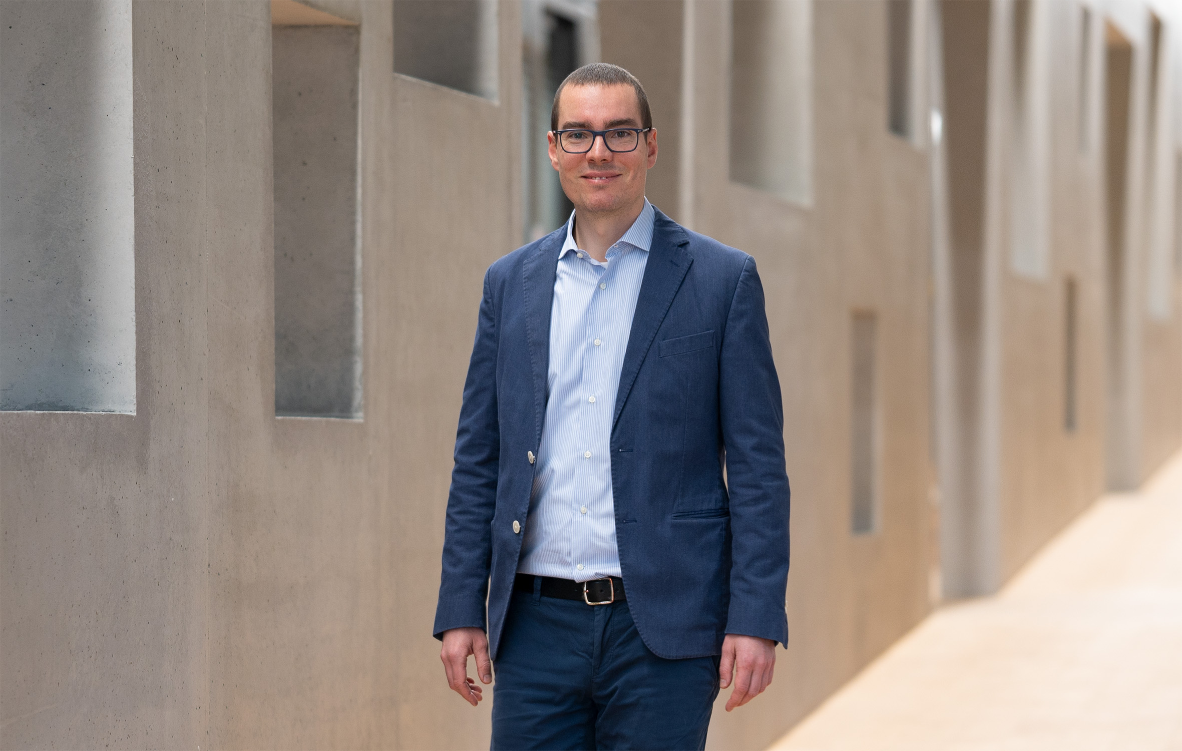 Ein Mann mit Brille, hellblauem Hemd und dunkelblauem Blazer steht selbstbewusst in einem Flur der Hochschule Coburg, umgeben von modernen Architekturelementen mit eckigen Formen und neutralen Tönen.