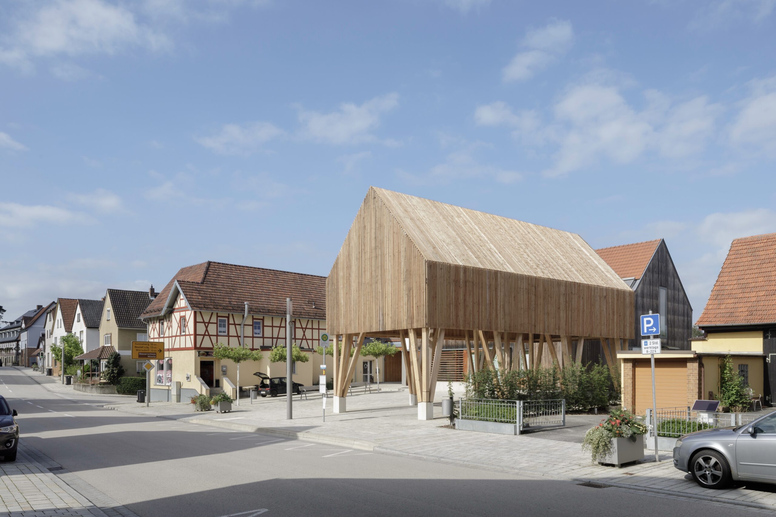 A street near Hochschule Coburg showcases traditional half-timbered houses alongside a modern open wooden structure on stilts. The partly cloudy sky and a few parked cars complete the scene, highlighting the harmonious blend of old and new architecture.