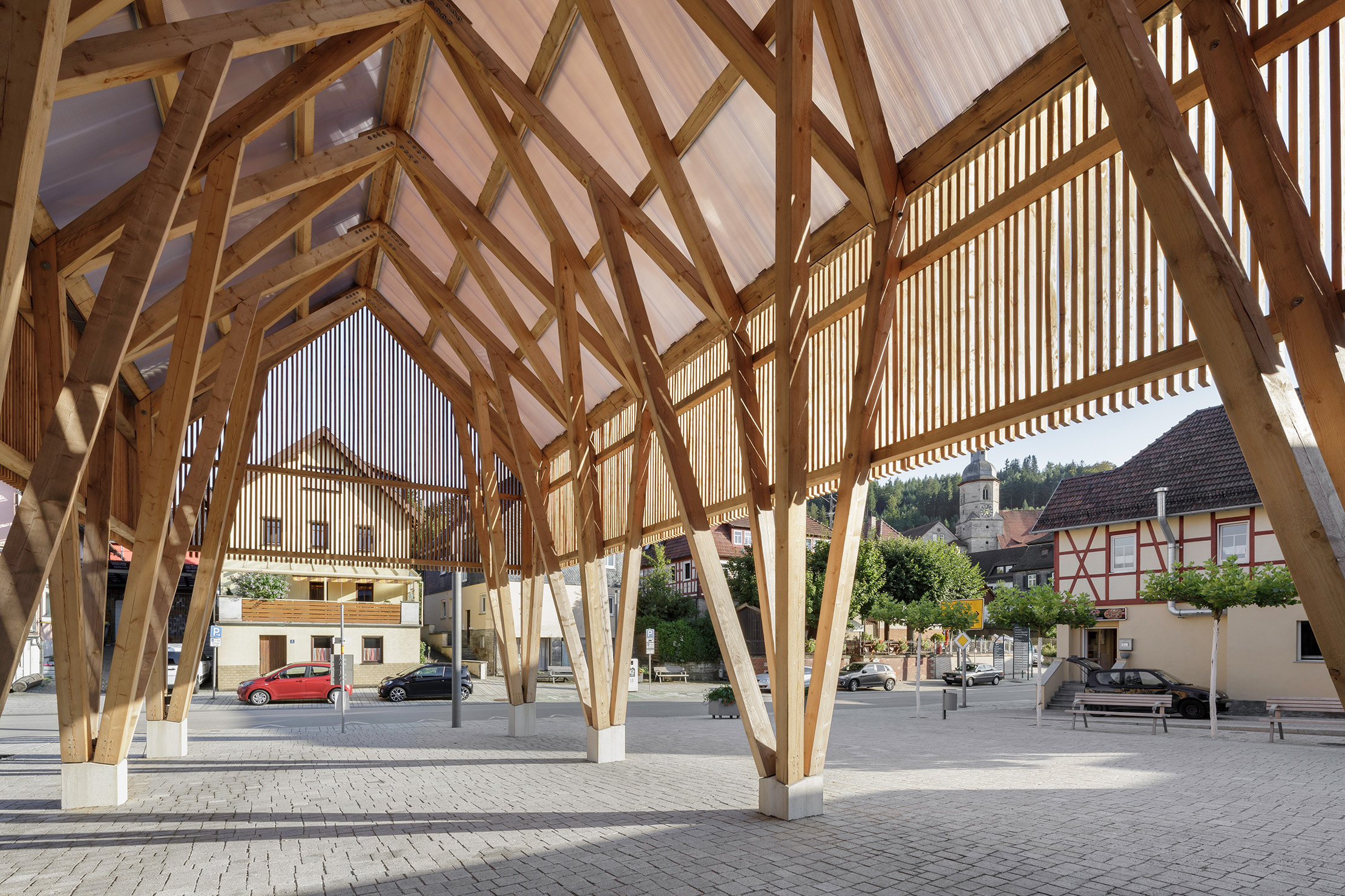 A modern wooden pergola with geometric beams provides shade over a cobblestone area at Hochschule Coburg. Traditional half-timbered houses surround the open space, with trees and a clear sky in the background.