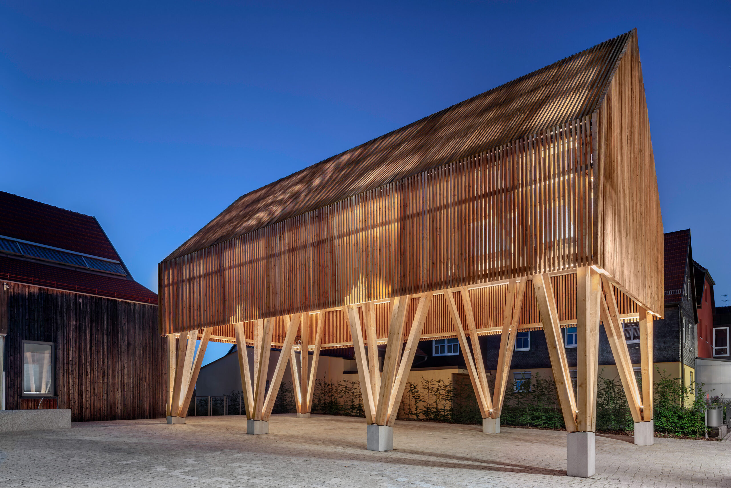 A modern wooden building elevated on stilts, featuring a slatted design with warm lighting, echoes the architectural aesthetics of Hochschule Coburg. It stands against a clear evening sky, surrounded by a simple paved area and neighboring structures.