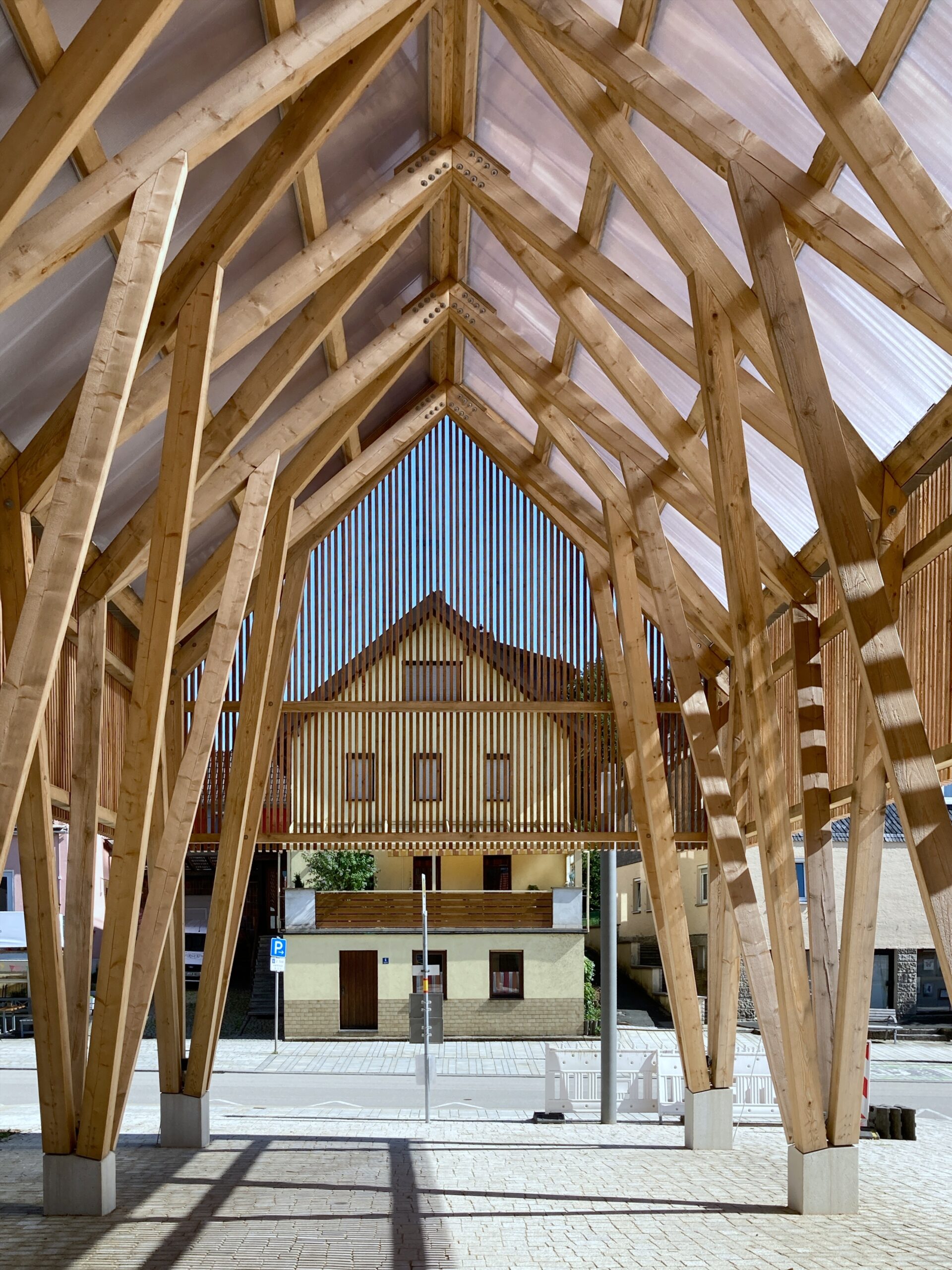 A wooden architectural structure with angled beams and a translucent roof creates a walkway view, framing Hochschule Coburg's traditional house with red roof tiles in the background. The scene is set on a sunny day under a clear blue sky.