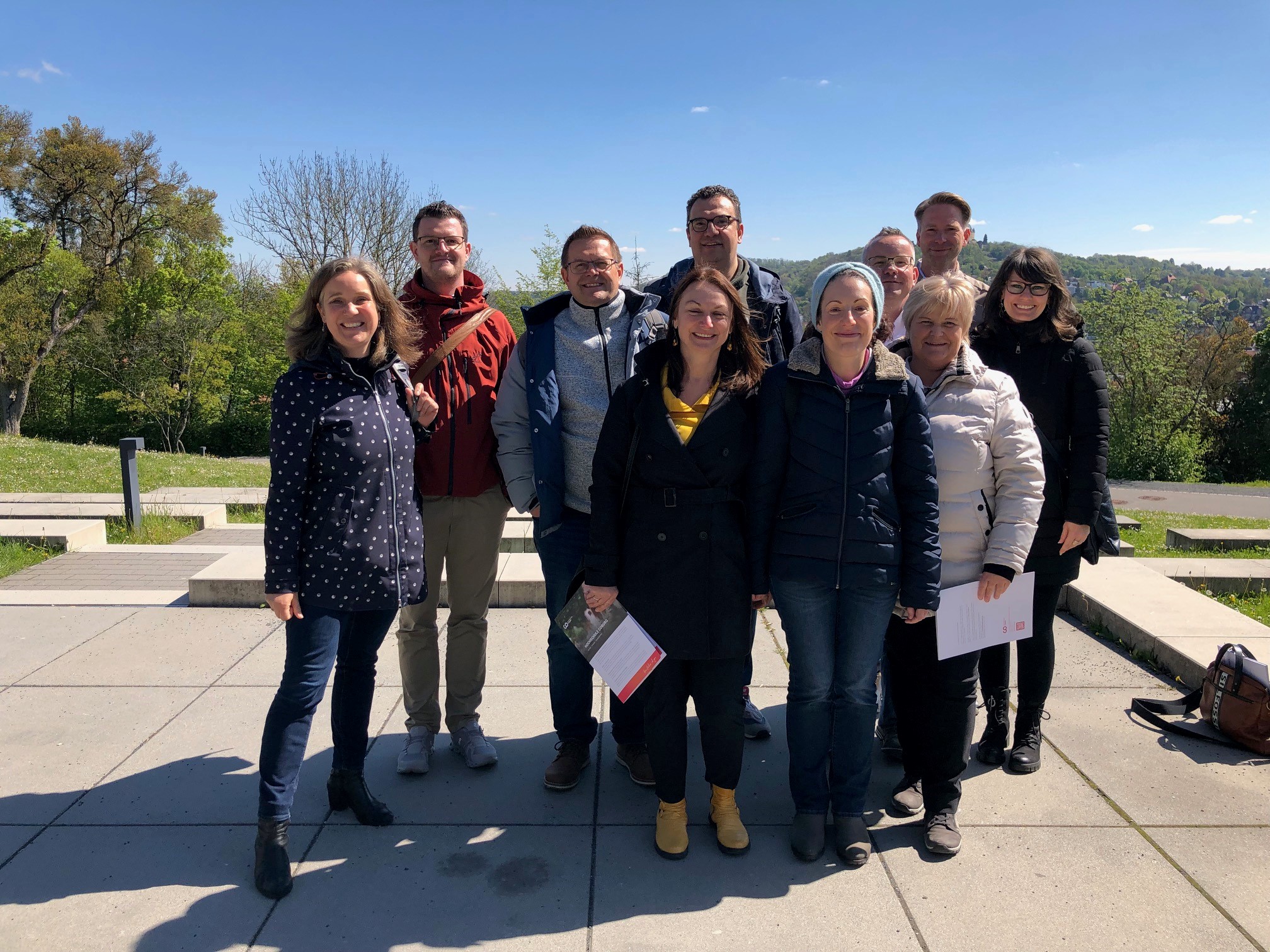 A group of people smiling outdoors on a sunny day at Hochschule Coburg, standing on a paved area surrounded by greenery. They are wearing jackets and holding papers, with a clear blue sky in the background.