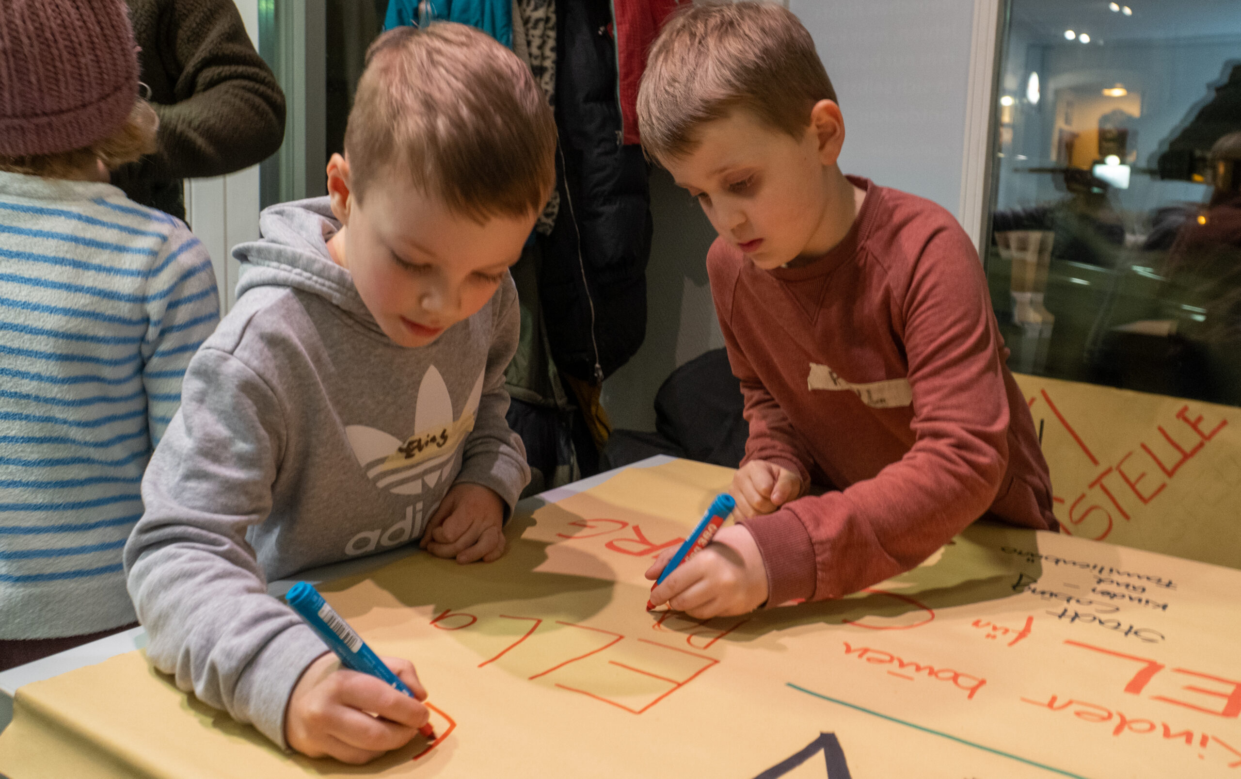 Zwei kleine Kinder der Hochschule Coburg schreiben mit Markern auf ein großes Blatt Papier. Sie konzentrieren sich auf ihre Aufgabe, umgeben von Mänteln und anderen Menschen in einer ungezwungenen Innenumgebung. Das Papier ist voller bunter Wörter und lebendiger Zeichnungen.