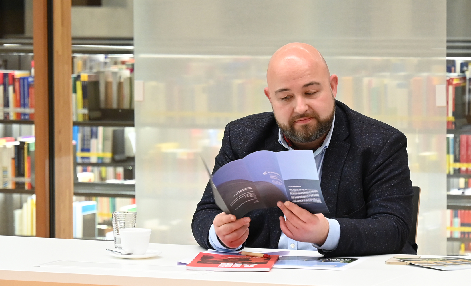 In einer ruhigen Ecke der Bibliothek der Hochschule Coburg sitzt ein kahlköpfiger Mann mit Bart an seinem Schreibtisch und liest vertieft ein blaues Büchlein. Er ist von Bücherregalen umgeben, während eine Tasse und Untertasse in Reichweite stehen und seine wissenschaftlichen Bestrebungen antreiben.