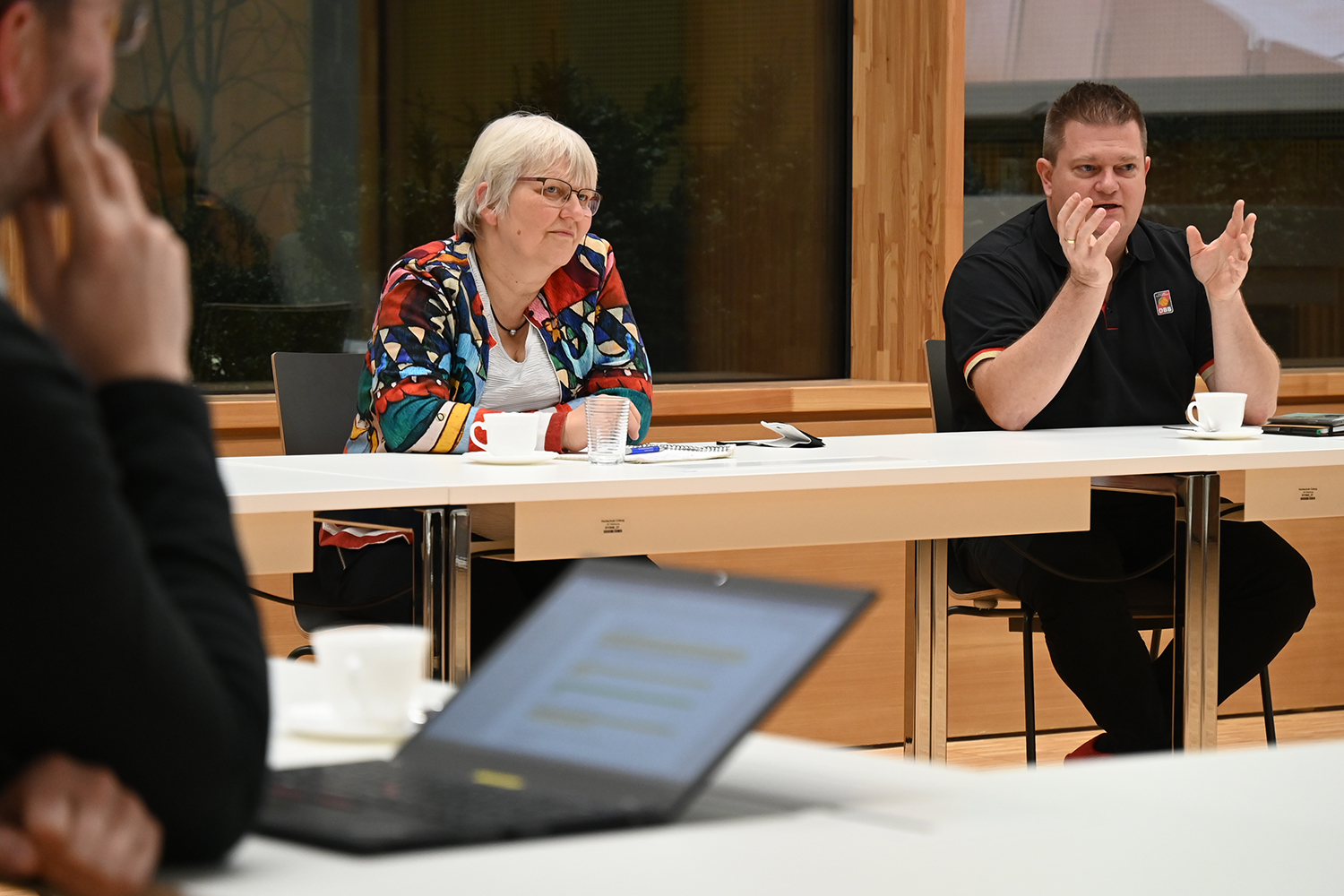 In einem Besprechungsraum der Hochschule Coburg sitzt eine Gruppe um einen Tisch. Eine Frau mit Brille und bunter Jacke unterhält sich mit einem Mann im schwarzen Hemd. Laptops und Kaffeetassen liegen verstreut auf dem Tisch, doch der Fokus liegt auf ihrem spannenden Dialog.