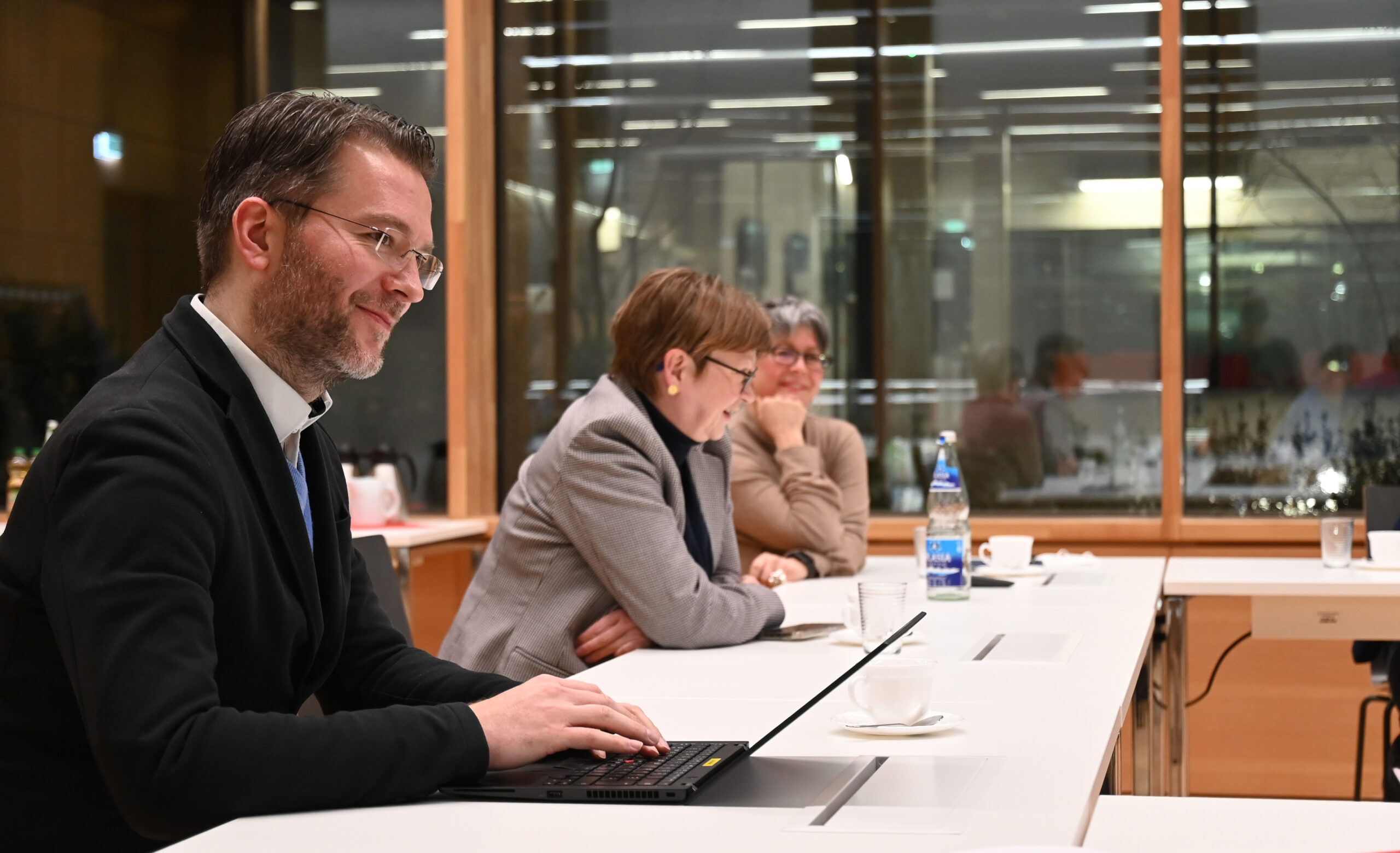 Ein Mann im Anzug arbeitet an einem Laptop und sitzt mit zwei Frauen von der Hochschule Coburg an einem Konferenztisch. Die Frauen unterhalten sich. Der Raum hat große Fenster mit Blick auf die Lichter der Stadt. Auf dem Tisch stehen Tassen und eine Wasserflasche.