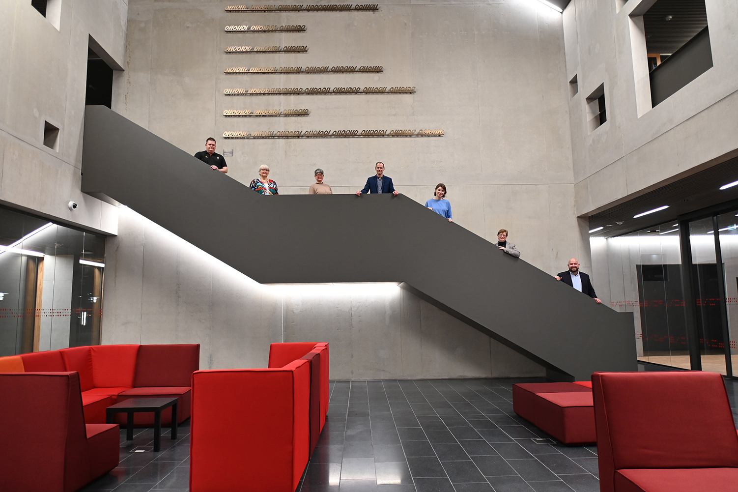 Eine Gruppe von sieben Personen steht auf einer eleganten grauen Treppe in einem modernen Gebäude der Hochschule Coburg mit Betonwänden. Die untere Ebene ist mit roten Sitzgelegenheiten auf einem dunklen Fliesenboden ausgestattet, während an der Wand über der Gruppe horizontale Leuchten angebracht sind.