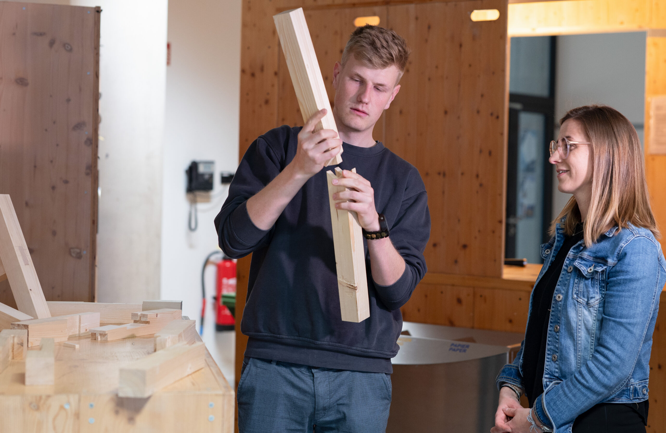 Ein Mann untersucht ein Holzbrett in einer Werkstatt der Hochschule Coburg, während eine Frau zusieht. Sie sind umgeben von komplizierten Holzkonstruktionen und Werkzeugen, und eine Mischung aus natürlichem und künstlichem Licht wertet die Szene auf.