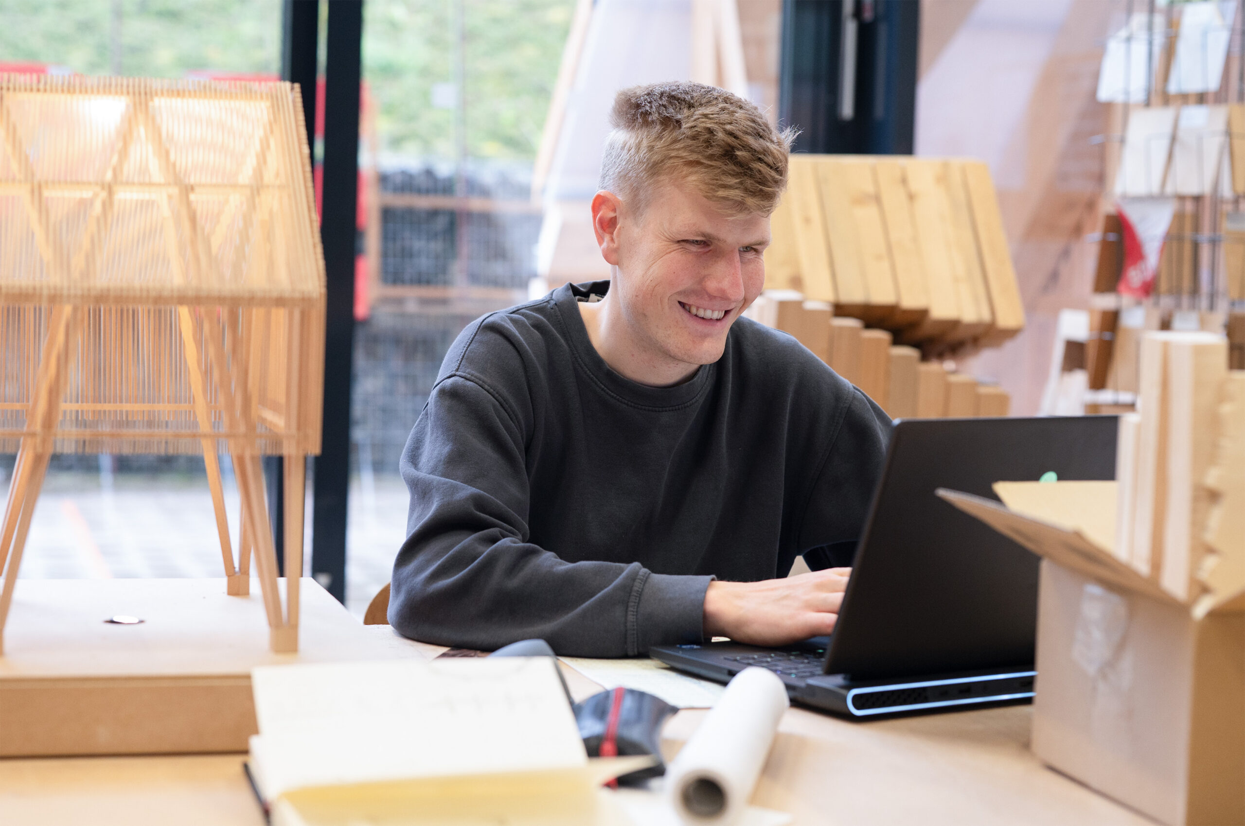 A person wearing a Hochschule Coburg dark sweatshirt is smiling while working on a laptop in a room filled with cardboard boxes and architectural models. The background features large windows with a blurred outdoor view.