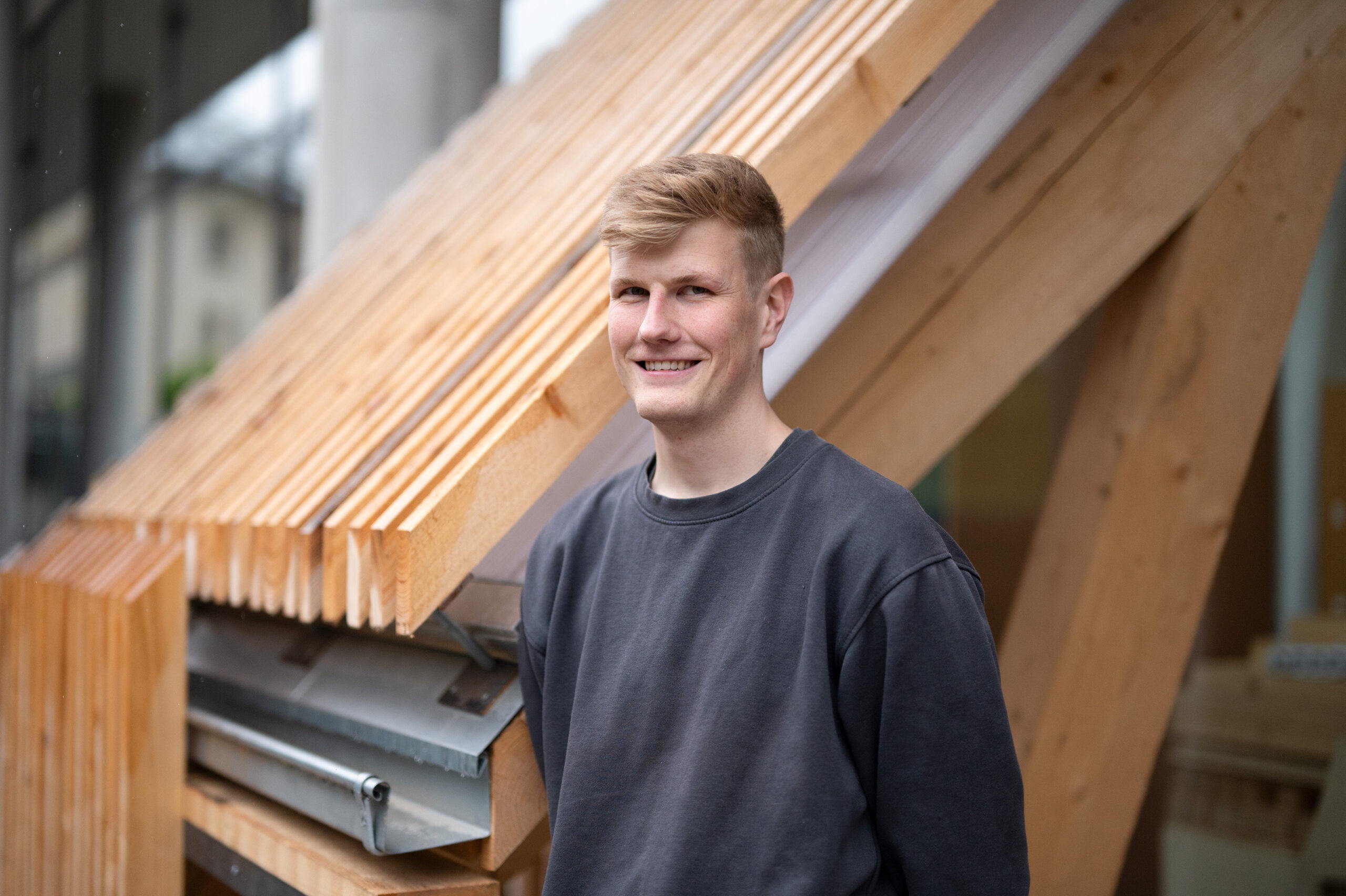 Eine Person mit kurzen Haaren und dunklem Sweatshirt steht lächelnd vor einer hölzernen Architekturstruktur mit überlappenden Balken. Die Szene, die an den innovativen Designstil der Hochschule Coburg erinnert, wird von verschwommenem Grün und einem angrenzenden Gebäude eingerahmt.