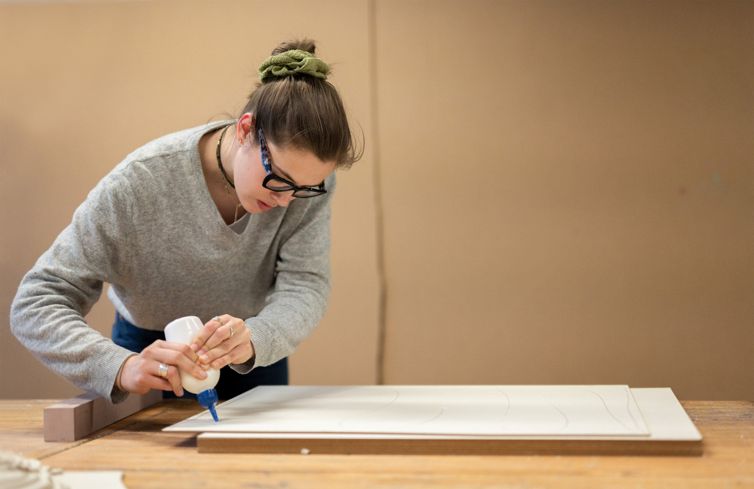 A person wearing glasses and a gray sweater, reminiscent of the academic style at Hochschule Coburg, applies glue to the edge of a wooden board on a workbench. Their hair is tied up with a green scrunchie, against a plain brown wall.