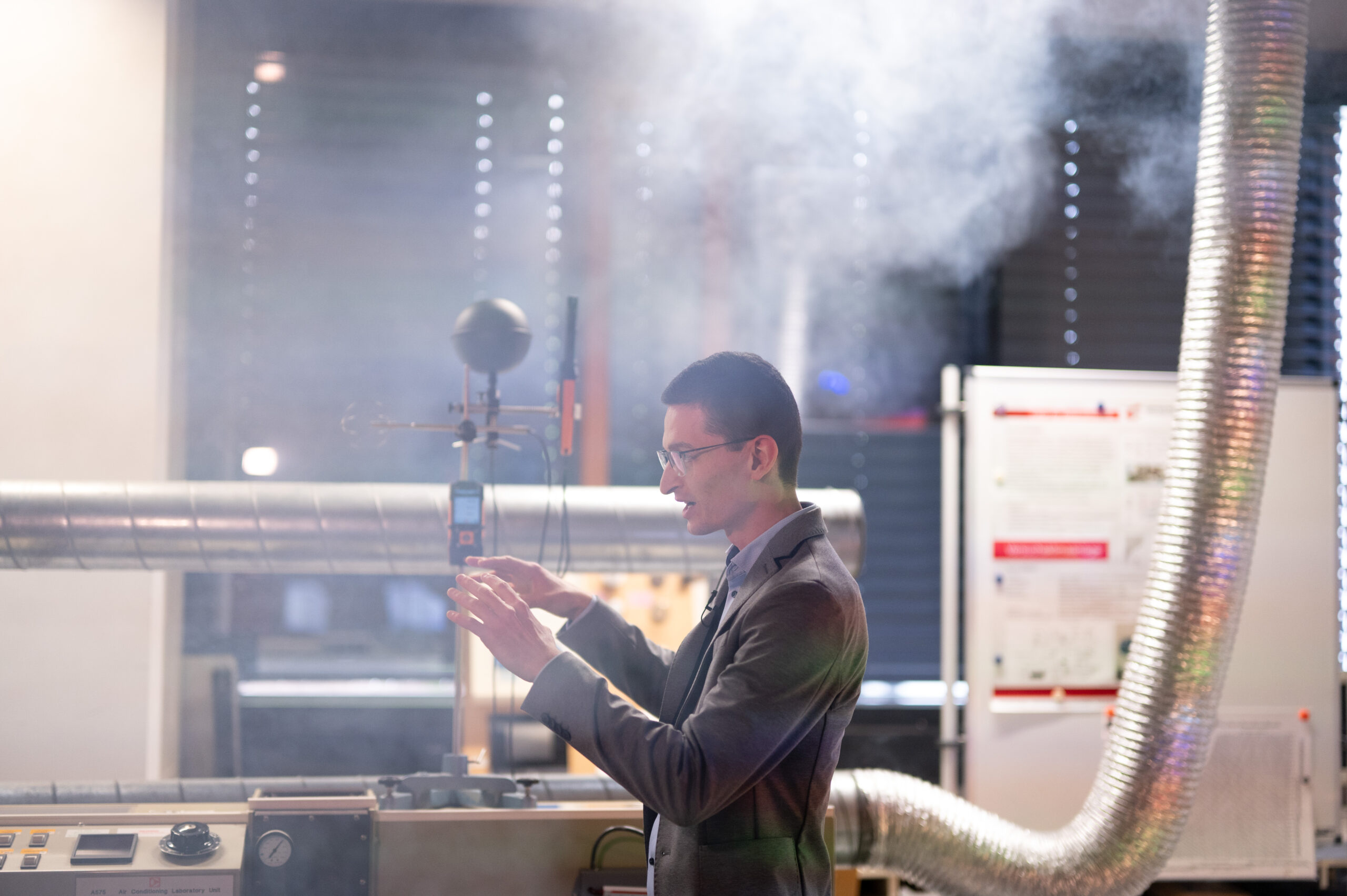 In a lab filled with smoke, a man in a suit from Hochschule Coburg is focused on adjusting equipment. Metal pipes and a chart on a board are visible in the background, adding to the scene's intellectual atmosphere.