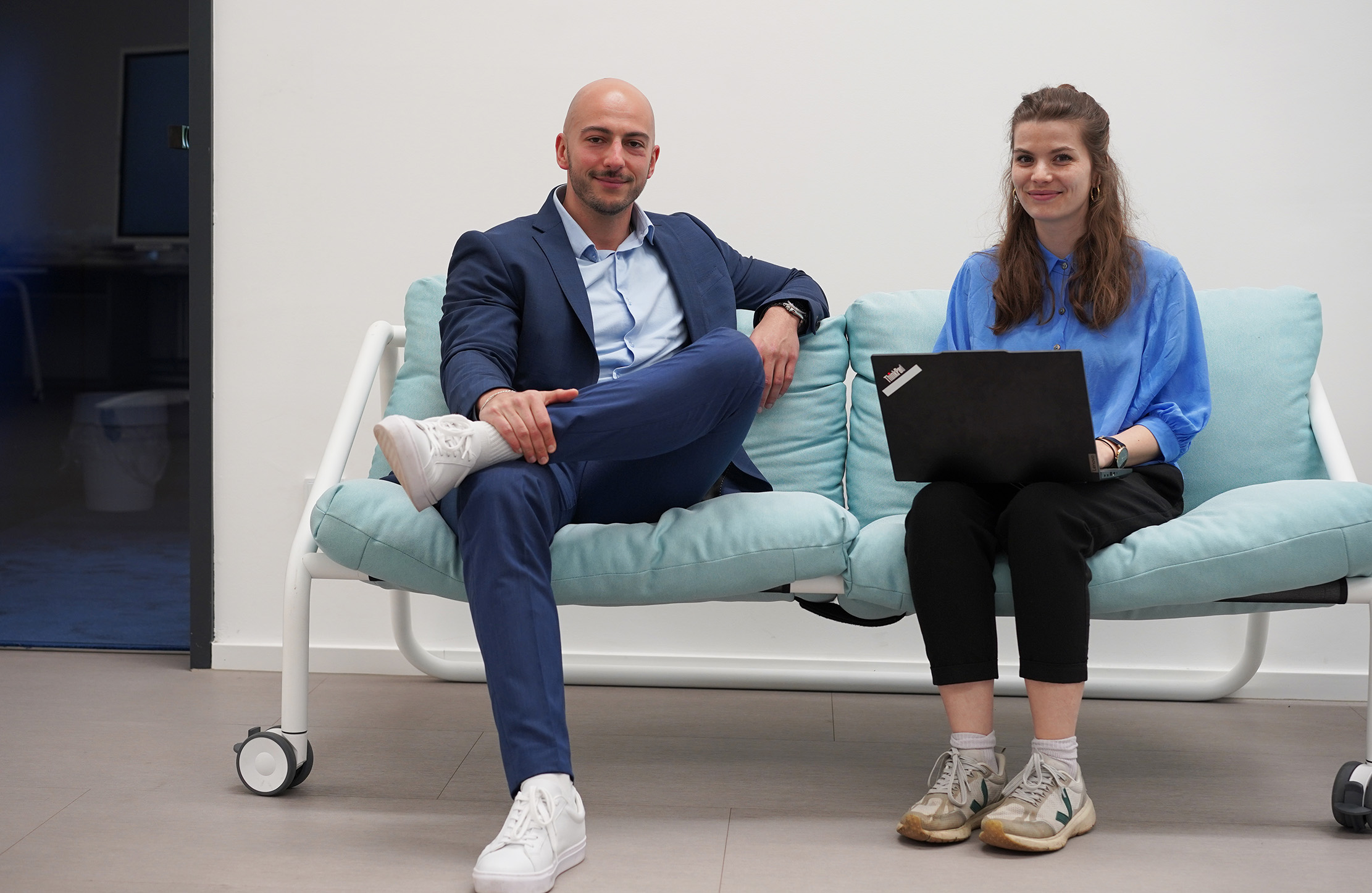 Ein Mann in einem blauen Anzug und weißen Turnschuhen sitzt auf einem hellblauen Sofa neben einer Frau in einem T-Shirt der Hochschule Coburg und schwarzen Hosen, die einen Laptop hält. Sie lächeln in einer Büroumgebung.