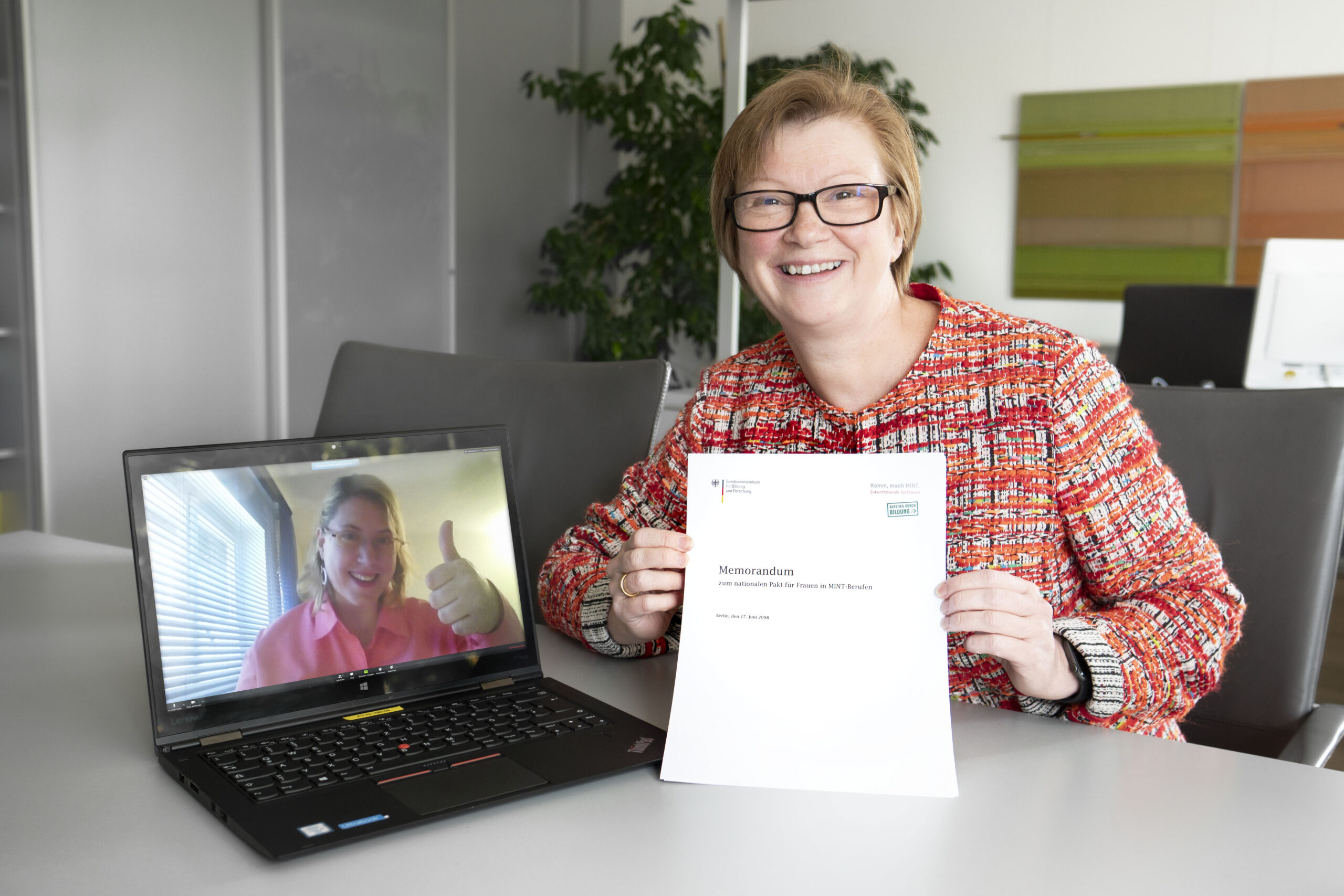 Eine Frau sitzt an einem Tisch und hält ein Dokument in der Hand. Sie lächelt in die Kamera. Auf ihrem Laptop läuft ein Videoanruf der Hochschule Coburg, eine andere Frau zeigt den Daumen nach oben. Im Hintergrund sind eine Pflanze und Aktenschränke zu sehen, die eine professionelle und zugleich einladende Atmosphäre schaffen.