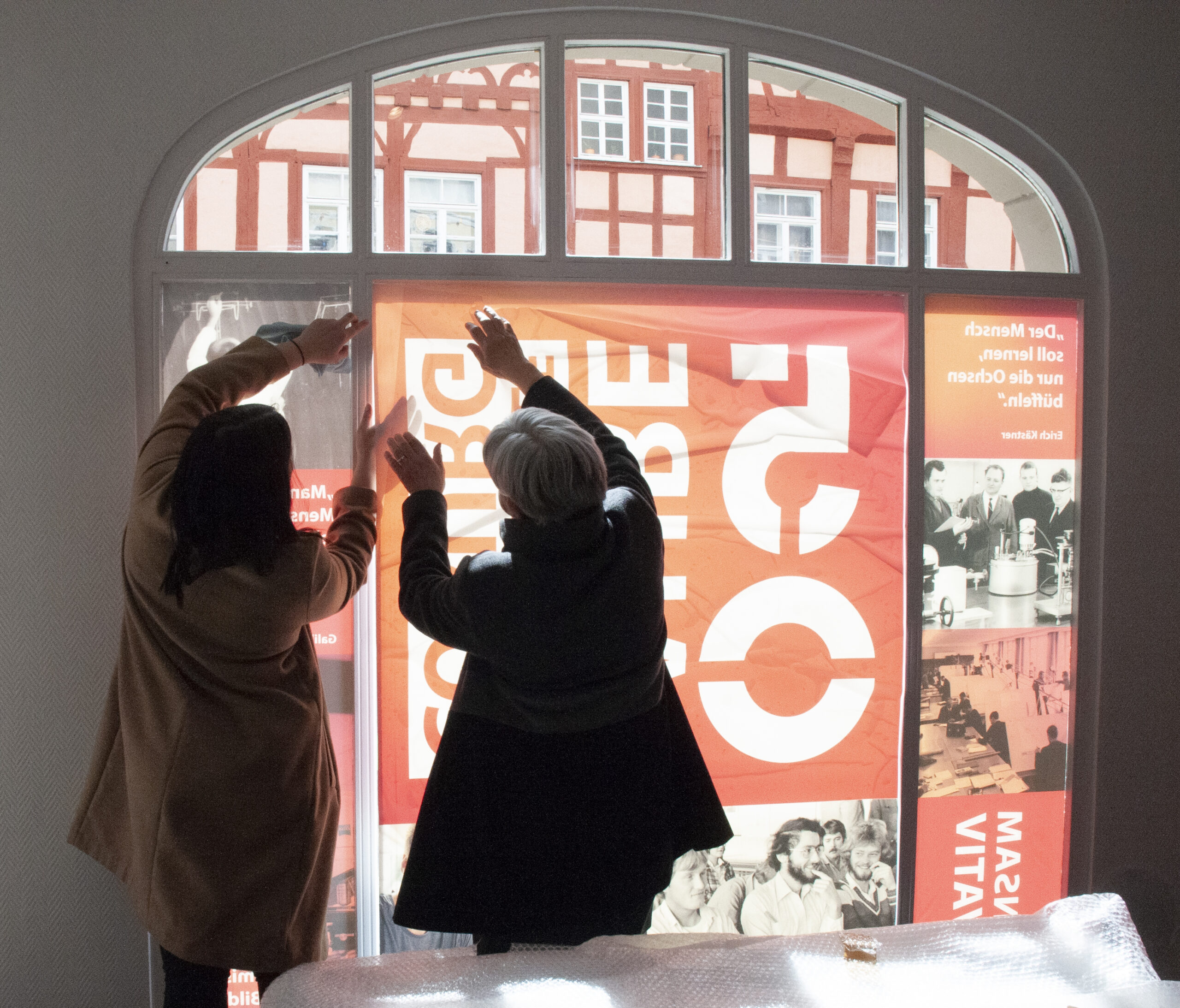 Zwei Personen hängen ein großes Plakat an ein Fenster der Hochschule Coburg. Das Plakat ist rot und weiß mit Text und Bildern. Sonnenlicht fällt durch das Glas und wirft Schatten auf ihre Hände, während das historische Gebäude draußen der Szene Charme verleiht.