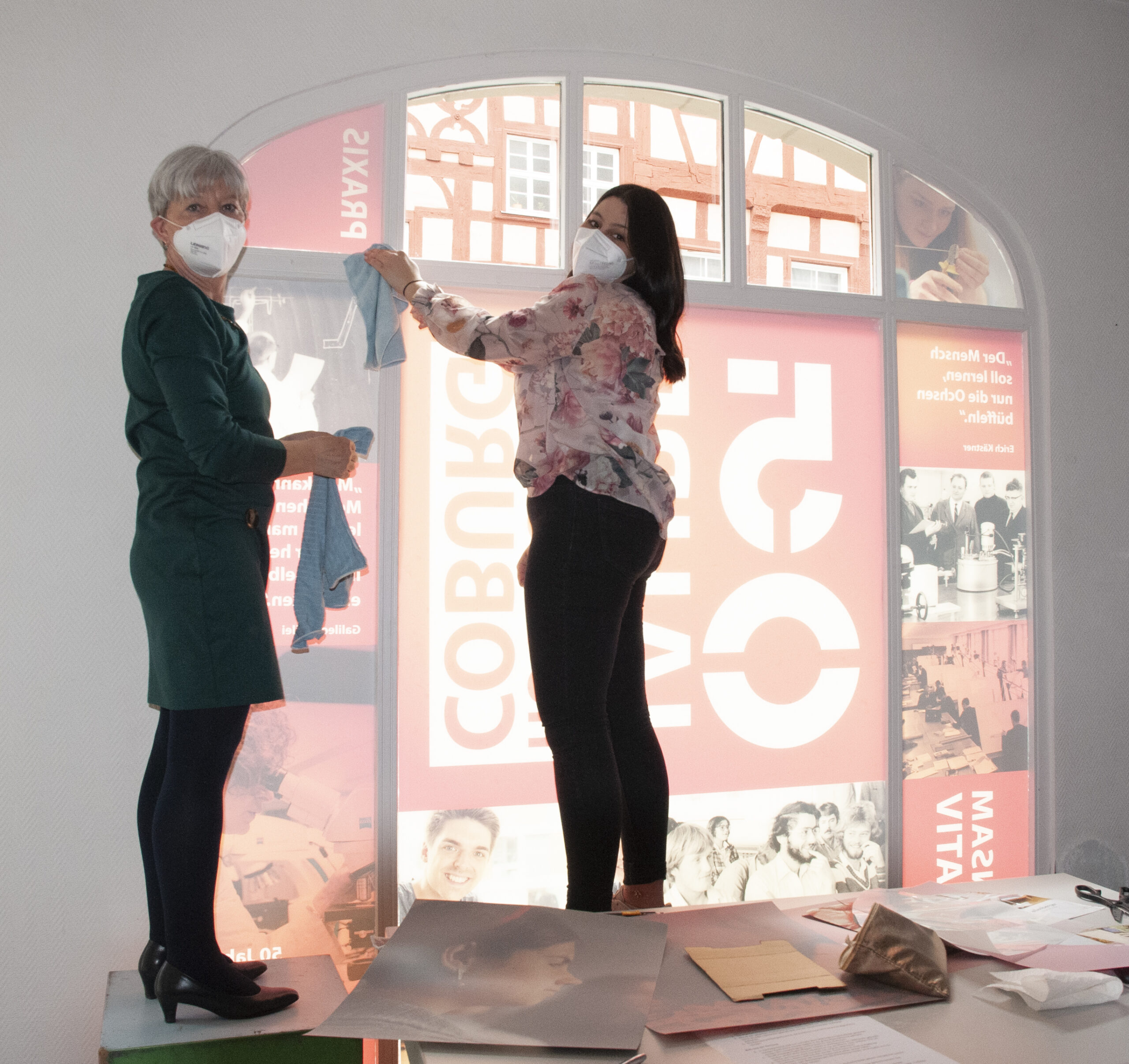 Zwei maskierte Personen stehen auf einem Tisch und hängen ein Tuch mit grafischen Motiven an ein großes Innenfenster der Hochschule Coburg. Die eine trägt ein grünes Kleid, die andere ein geblümtes Hemd und verleiht dem Arbeits- oder Ausstellungsbereich ein künstlerisches Flair.