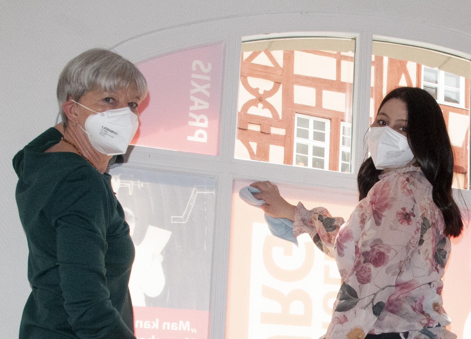 Zwei maskierte Personen stehen drinnen vor einem Fenster der Hochschule Coburg. Eine mit kurzen grauen Haaren ist grün gekleidet, während die andere mit langen dunklen Haaren eine geblümte Bluse trägt. Sie scheinen das Fenster zu putzen, das eine Szene im Freien widerspiegelt.