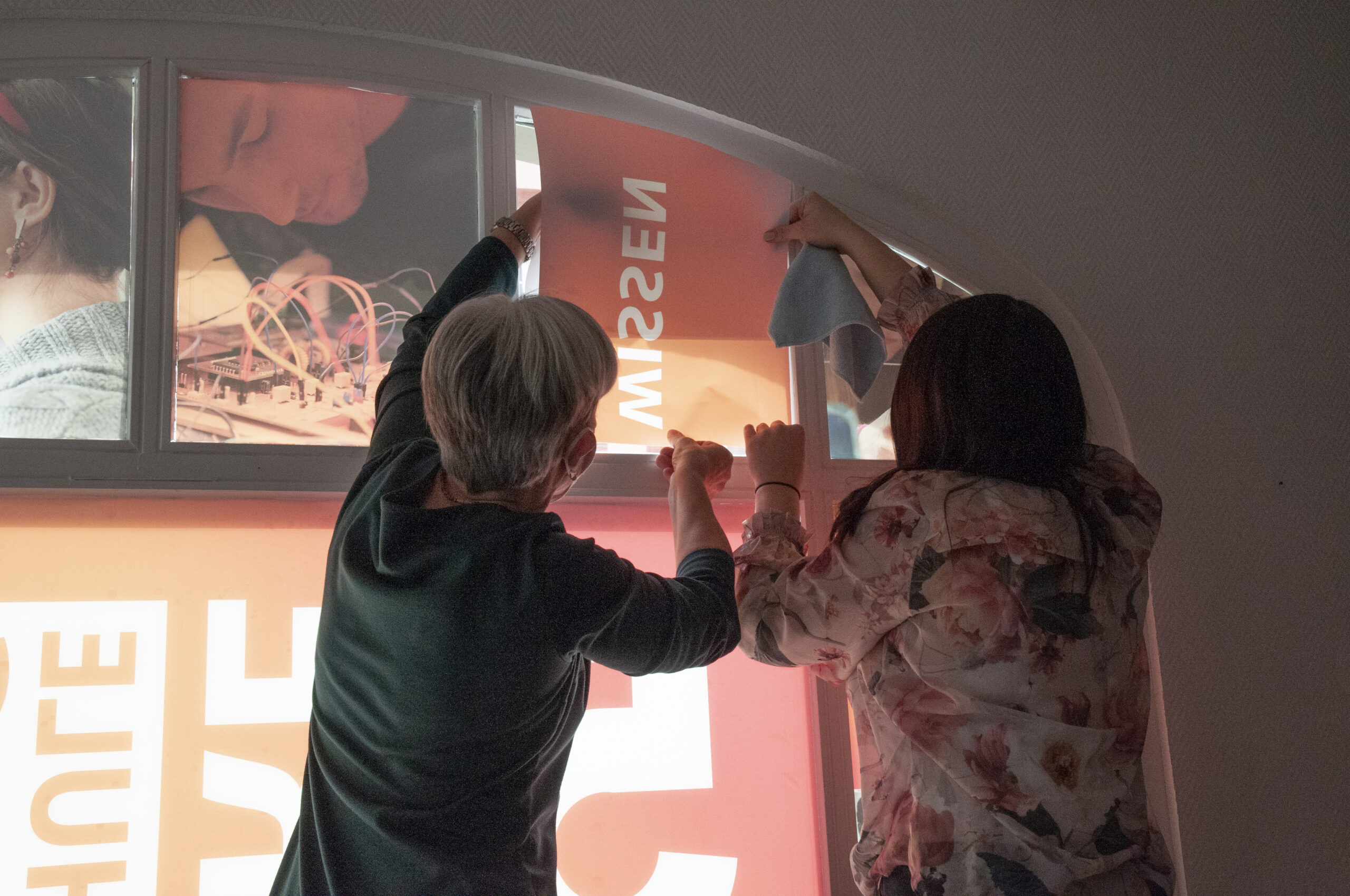 Zwei Personen befestigen ein großes orangefarbenes Plakat an einem Bogenfenster der Hochschule Coburg. Eine hält ein Tuch in der Hand, während sie das Plakat zurechtrückt. Helles Licht fällt durch das Plakat und beleuchtet den weißen Text und Teile der Bilder darauf. Die Szene spielt in einem schwach beleuchteten Raum.