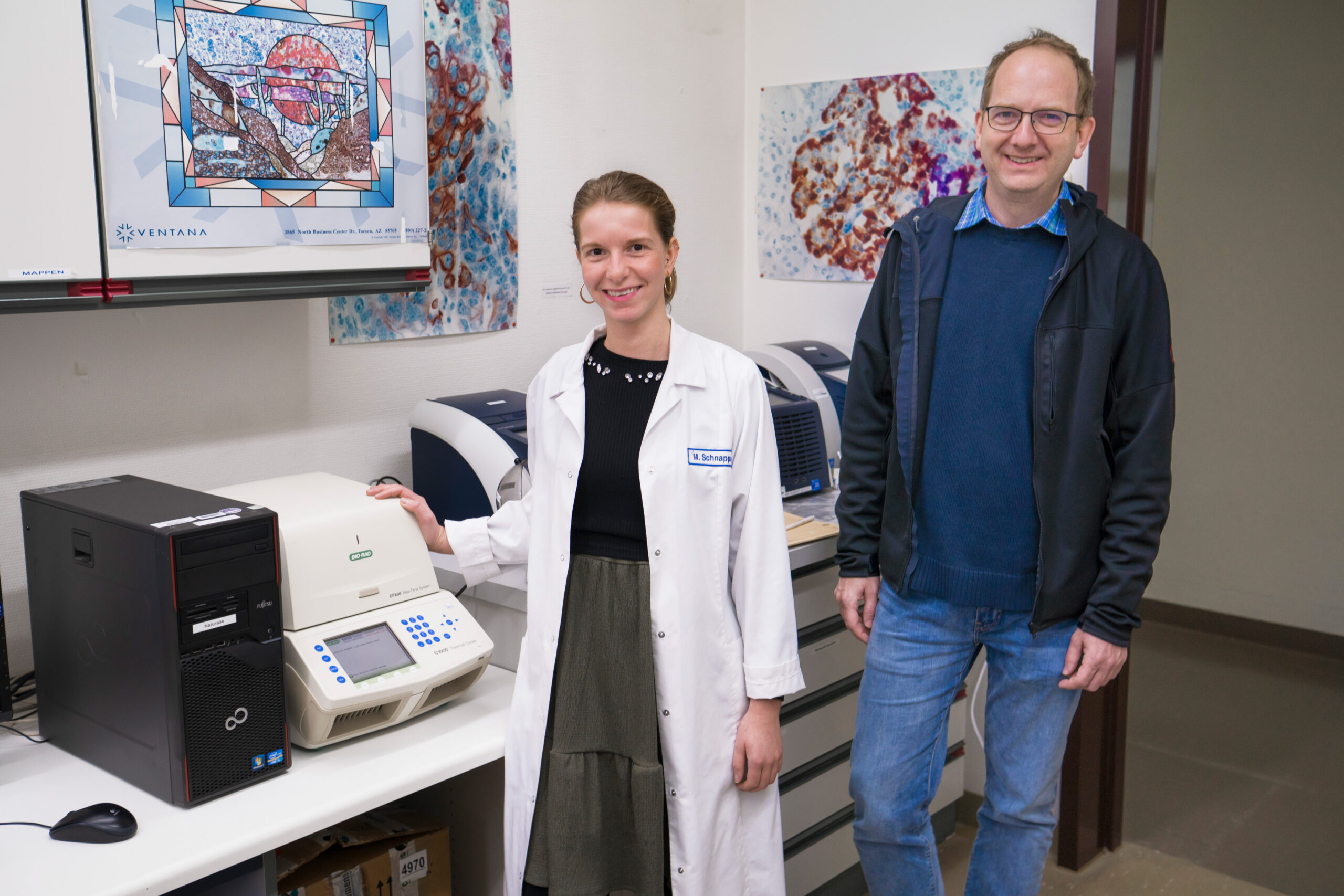 Eine Frau im Laborkittel, Vertreterin der Hochschule Coburg, steht lächelnd neben einer Labormaschine. Ein Mann mit Brille und Freizeitkleidung steht neben ihr. Dahinter hängen wissenschaftliche Bilder an der Wand und andere Laborgeräte, die eine lebendige Laboratmosphäre schaffen.