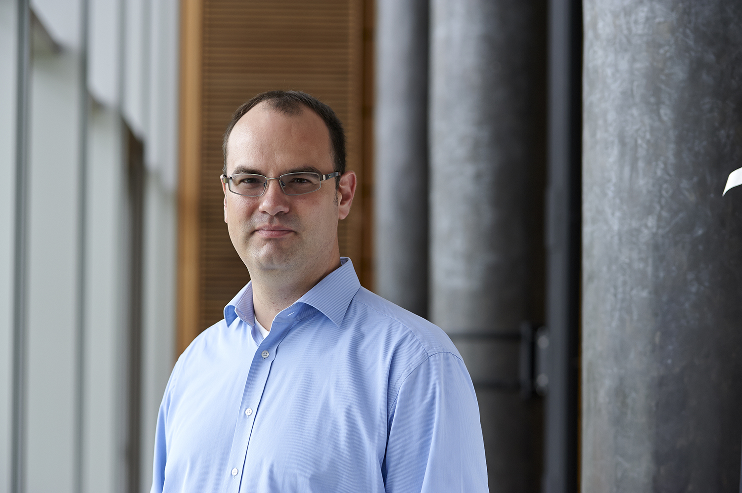 Ein Mann im hellblauen Hemd steht in einem modernen Flur der Hochschule Coburg, mit großen Fenstern links und zylindrischen Säulen rechts. Er hat kurze Haare und eine Brille und blickt mit neutralem Gesichtsausdruck in die Kamera.